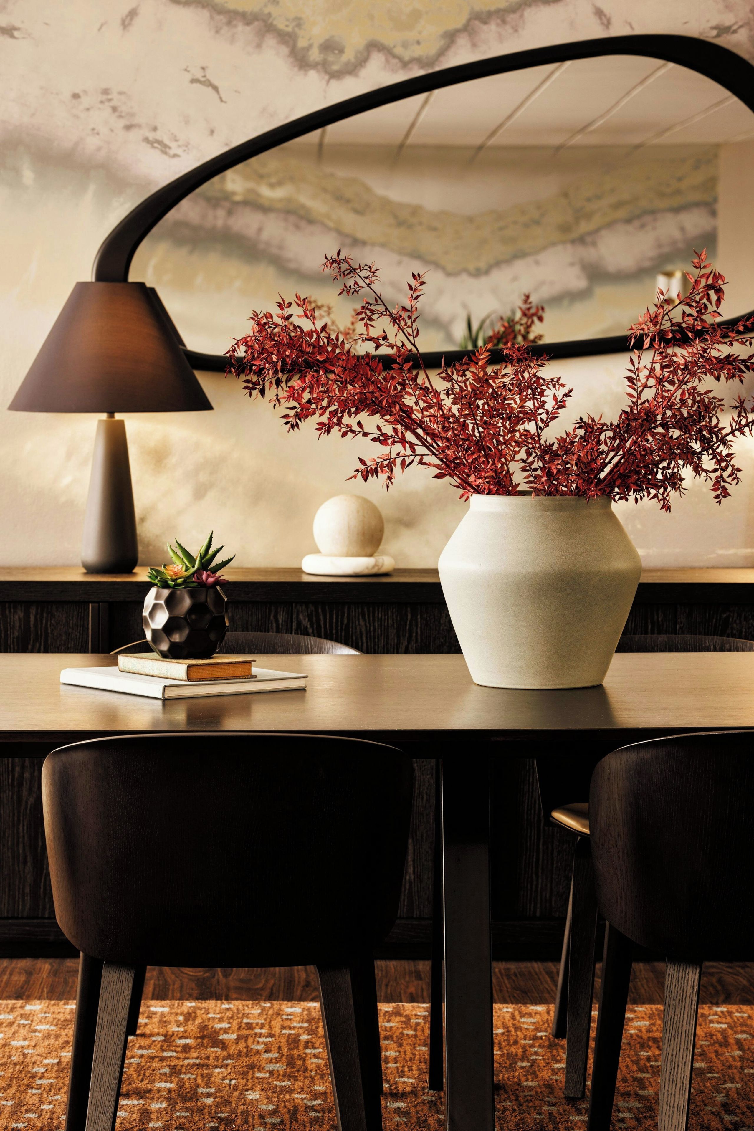 A dining area with a table, chairs, a lamp, a vase with red foliage, a pineapple, and a spherical object.