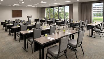 Un salón de conferencias con varias mesas y sillas, amplias ventanas con vista a espacios verdes y un letrero que indica 'The Westin'.
