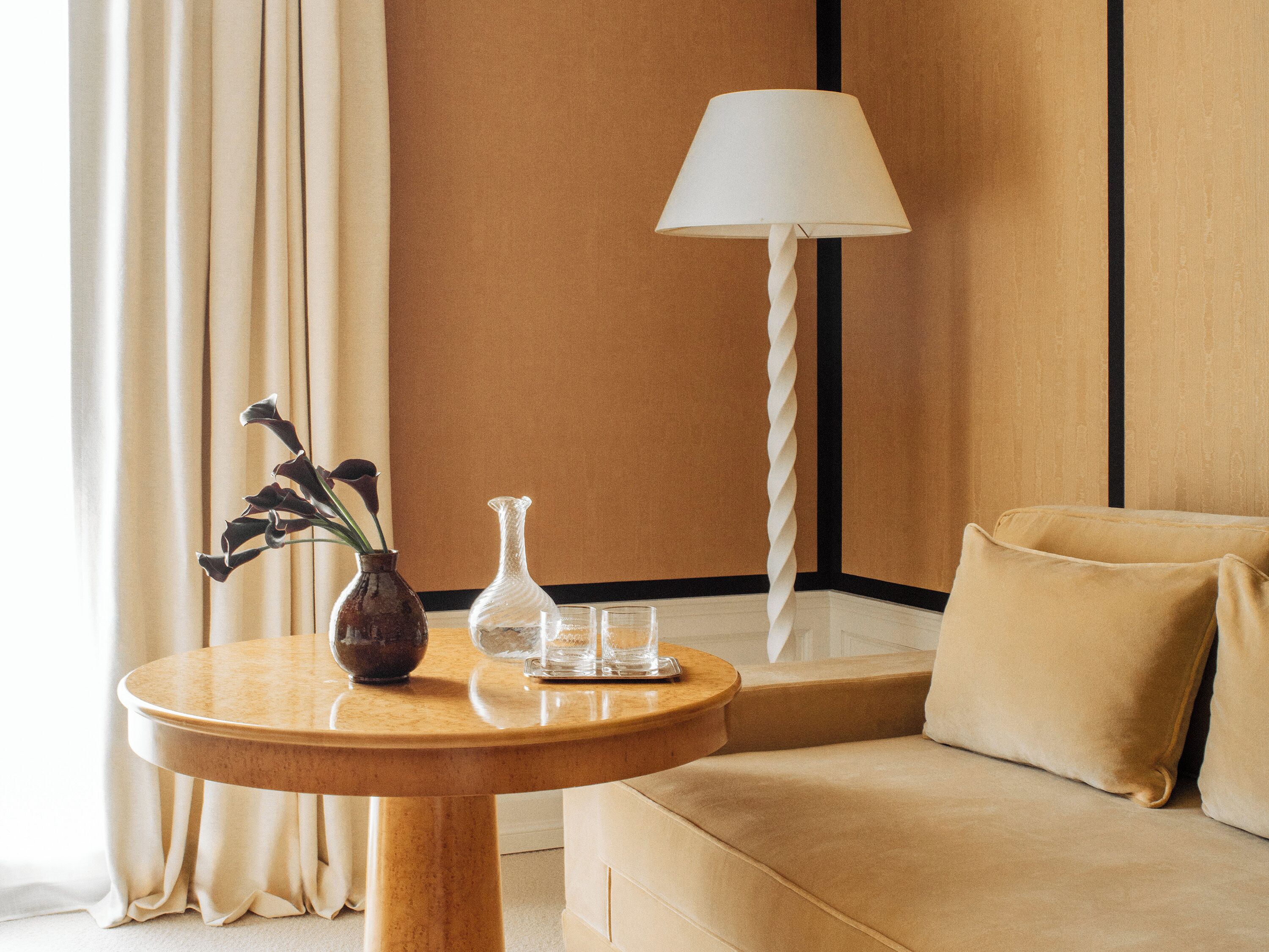 Una habitación con una mesa redonda de madera, un sofá, una lámpara, un jarrón con flores y una jofaina de vidrio.