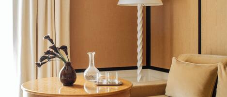 Una habitación con una mesa redonda de madera, un sofá, una lámpara, un jarrón con flores y una jofaina de vidrio.
