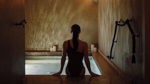 Una persona sentada junto a una piscina en un ambiente de spa, con suelo de madera y una pared cálida y con textura.