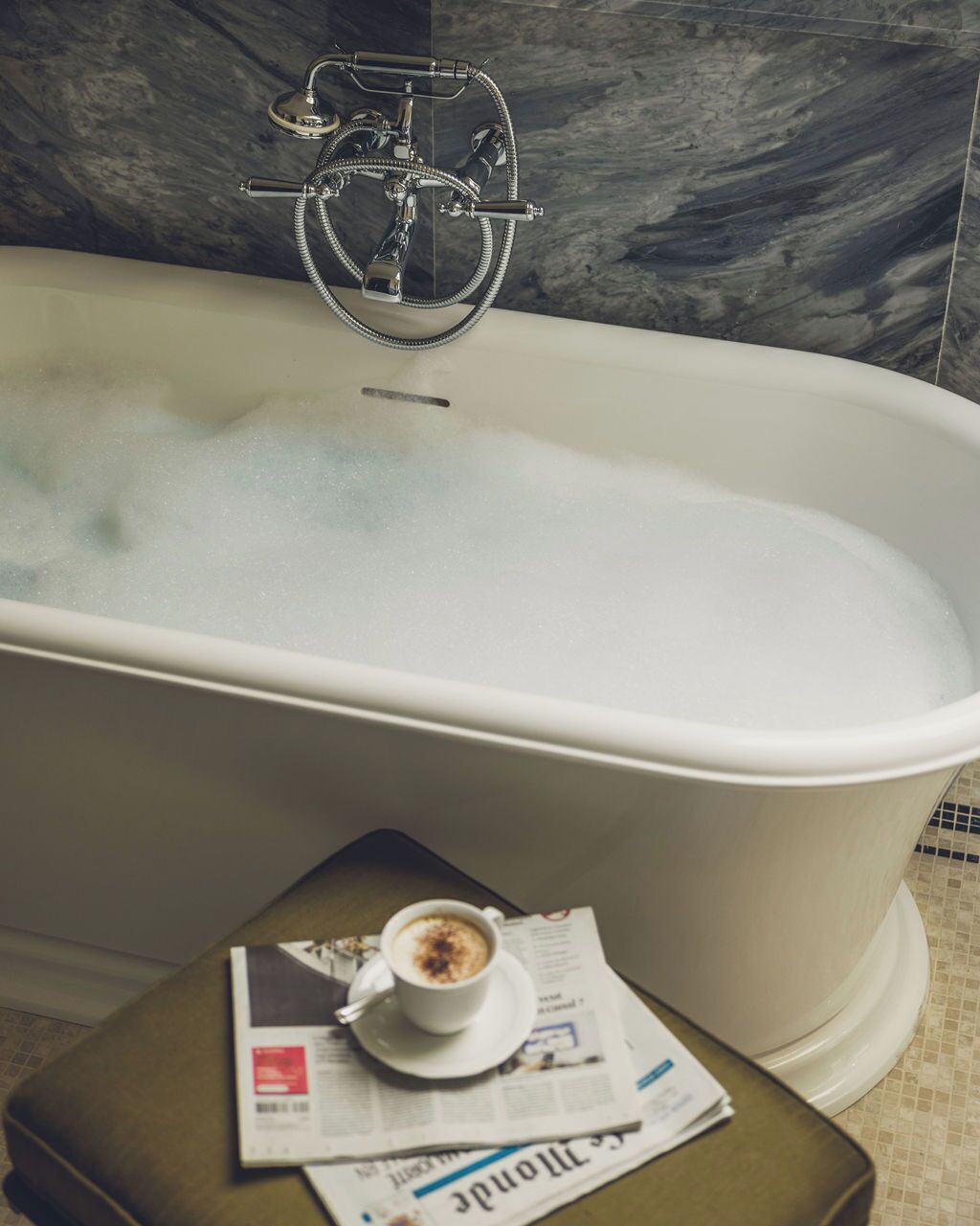 A bathtub with a showerhead, a cup of coffee on a newspaper, and a tiled floor.