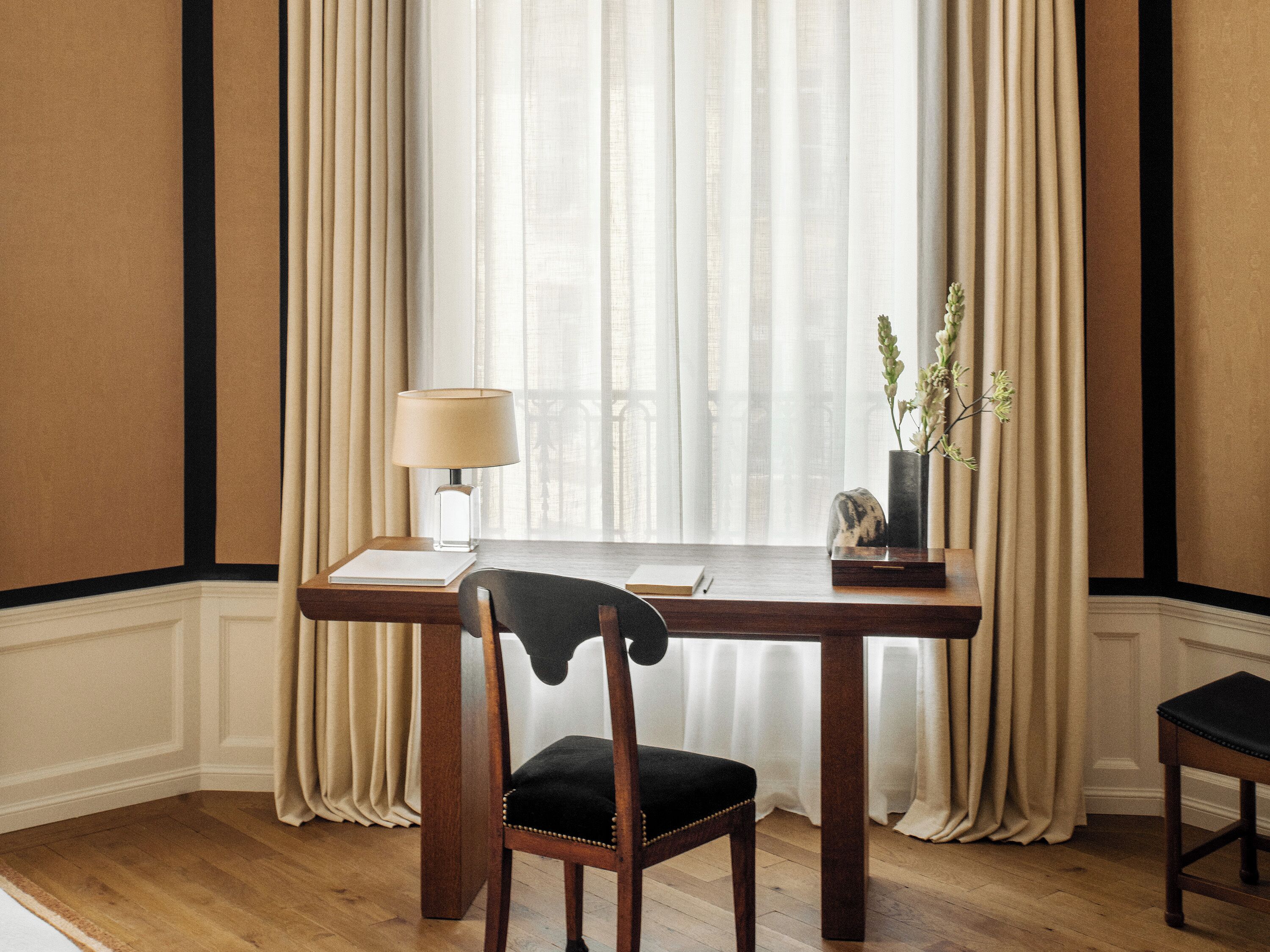 Ein Zimmer mit Holztisch, einem Stuhl, einer Lampe und einem Fenster mit durchsichtigen Vorhängen.