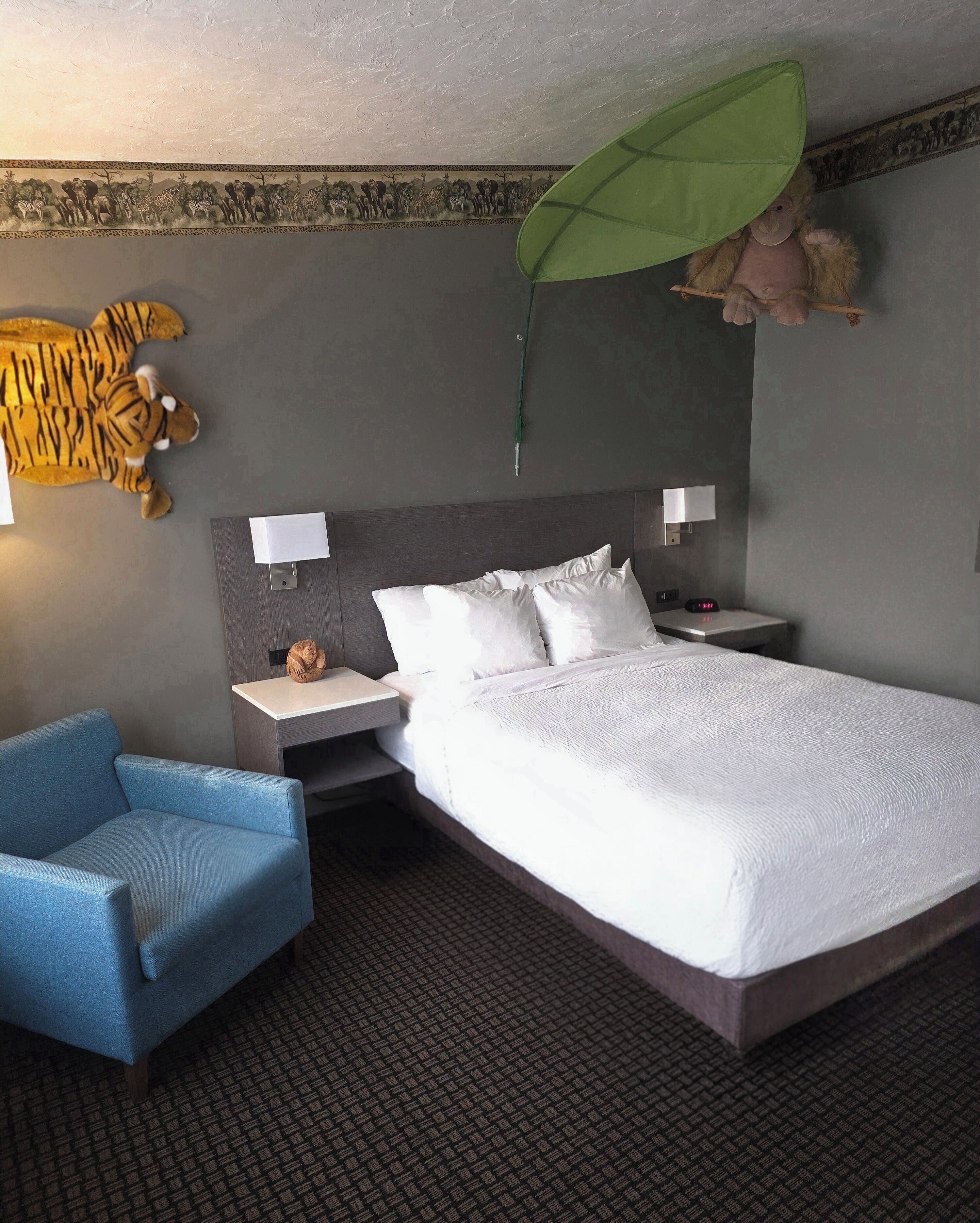 Una habitación de hotel con una cama grande, un sillón azul, una mesita de noche con una lámpara y un adorno en la pared.
