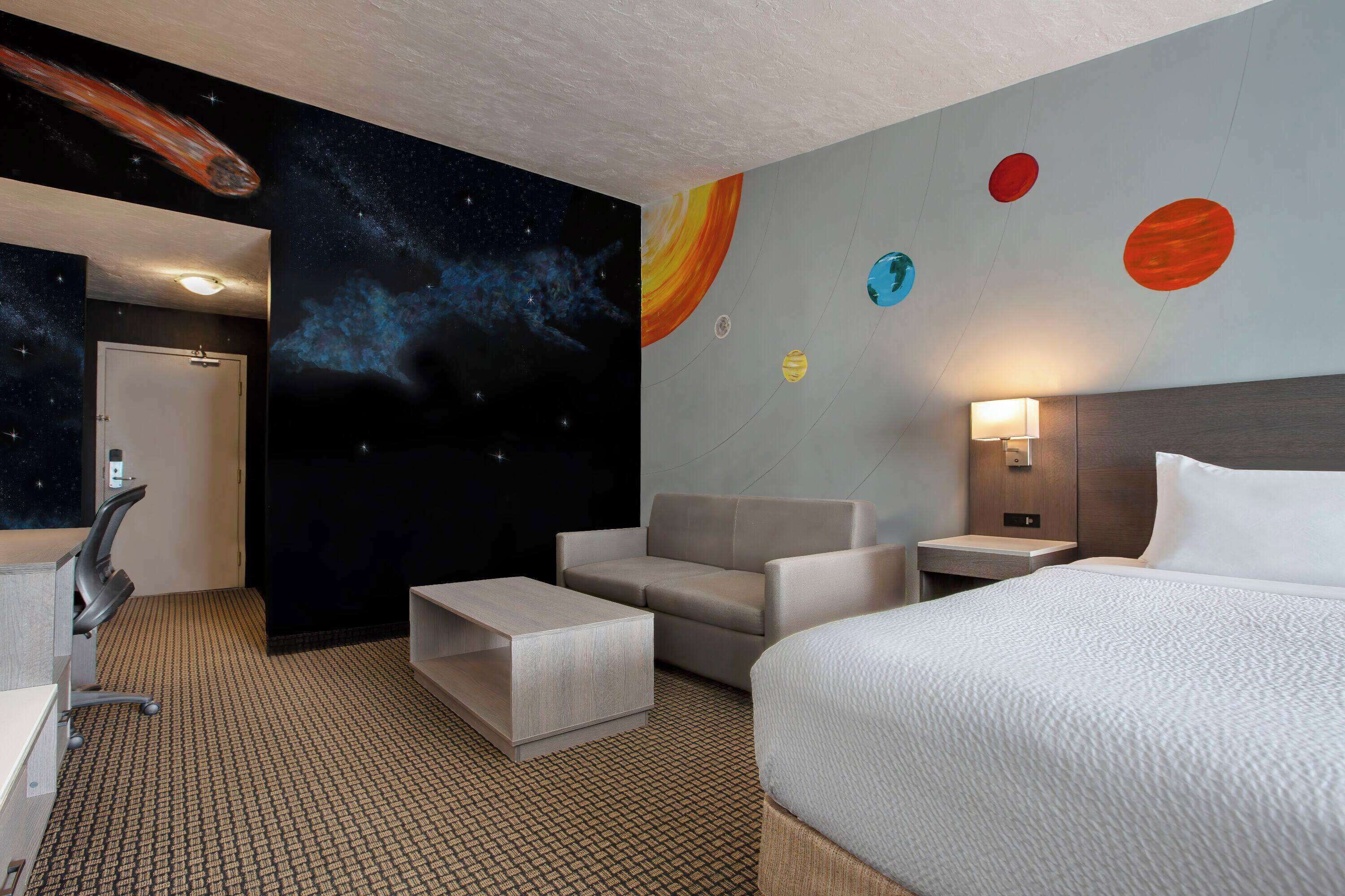 A hotel room with a bed, a sofa, a desk, and a wall with a space-themed mural.