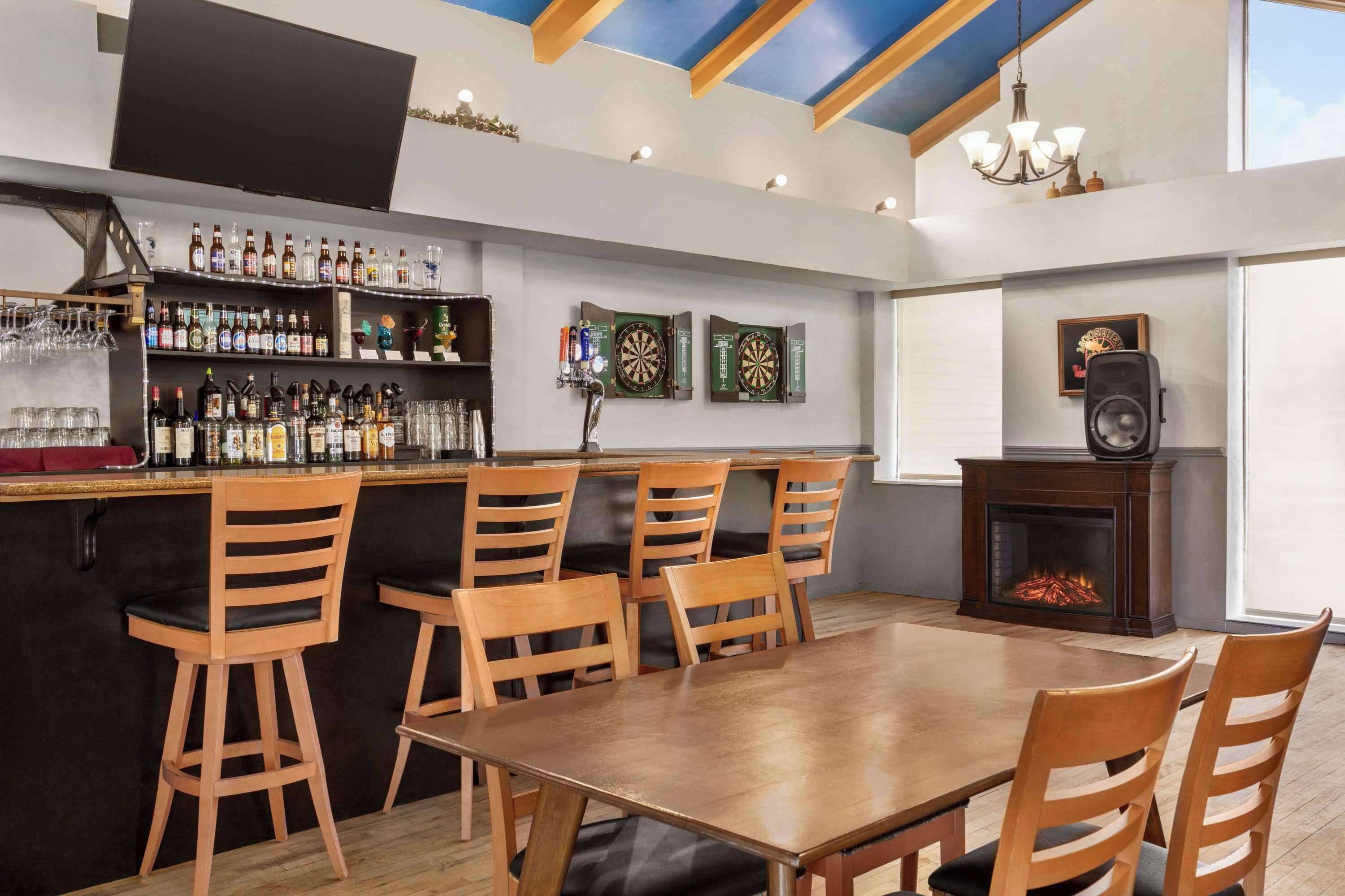 A bar area with wooden chairs and a table, shelves with bottles and glasses, dartboards, and a fireplace.