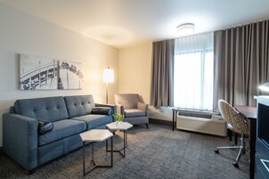Une chambre d’hôtel avec un canapé bleu, un fauteuil gris, une petite table avec une lampe, un bureau avec une chaise et une grande fenêtre avec des rideaux.
