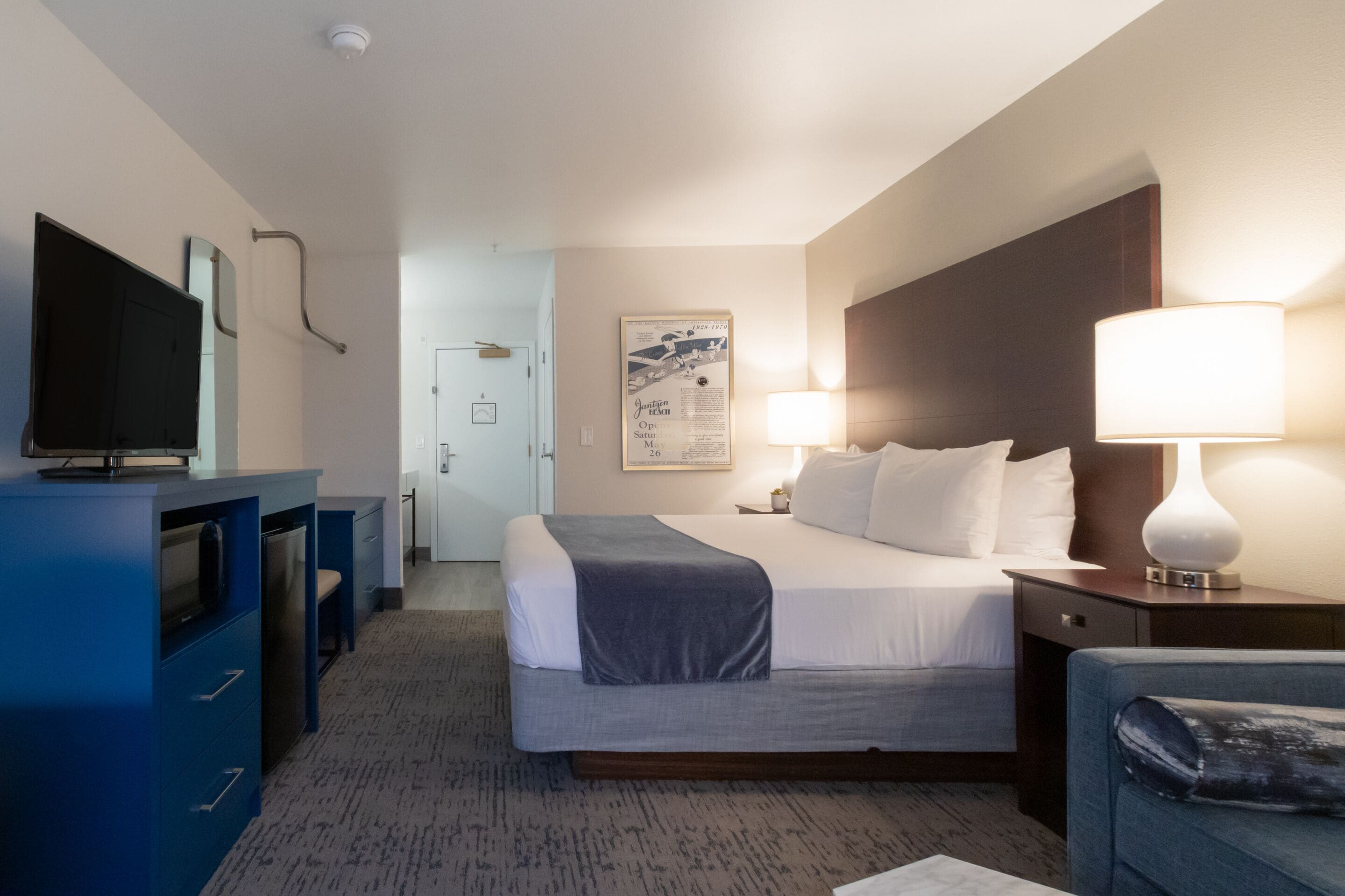 A hotel room with a bed, bedside table, lamp, TV, and a desk with drawers.
