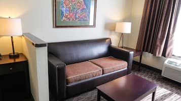 A hotel room with a leather sofa, a wooden coffee table, two bedside lamps, a framed artwork on the wall, and a window with curtains.