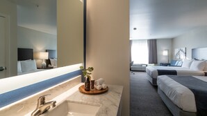 A hotel room with a marble bathroom sink, a large mirror, and a bed with a dark headboard. - Oxford Suites Portland - Jantzen Beach (Portland)