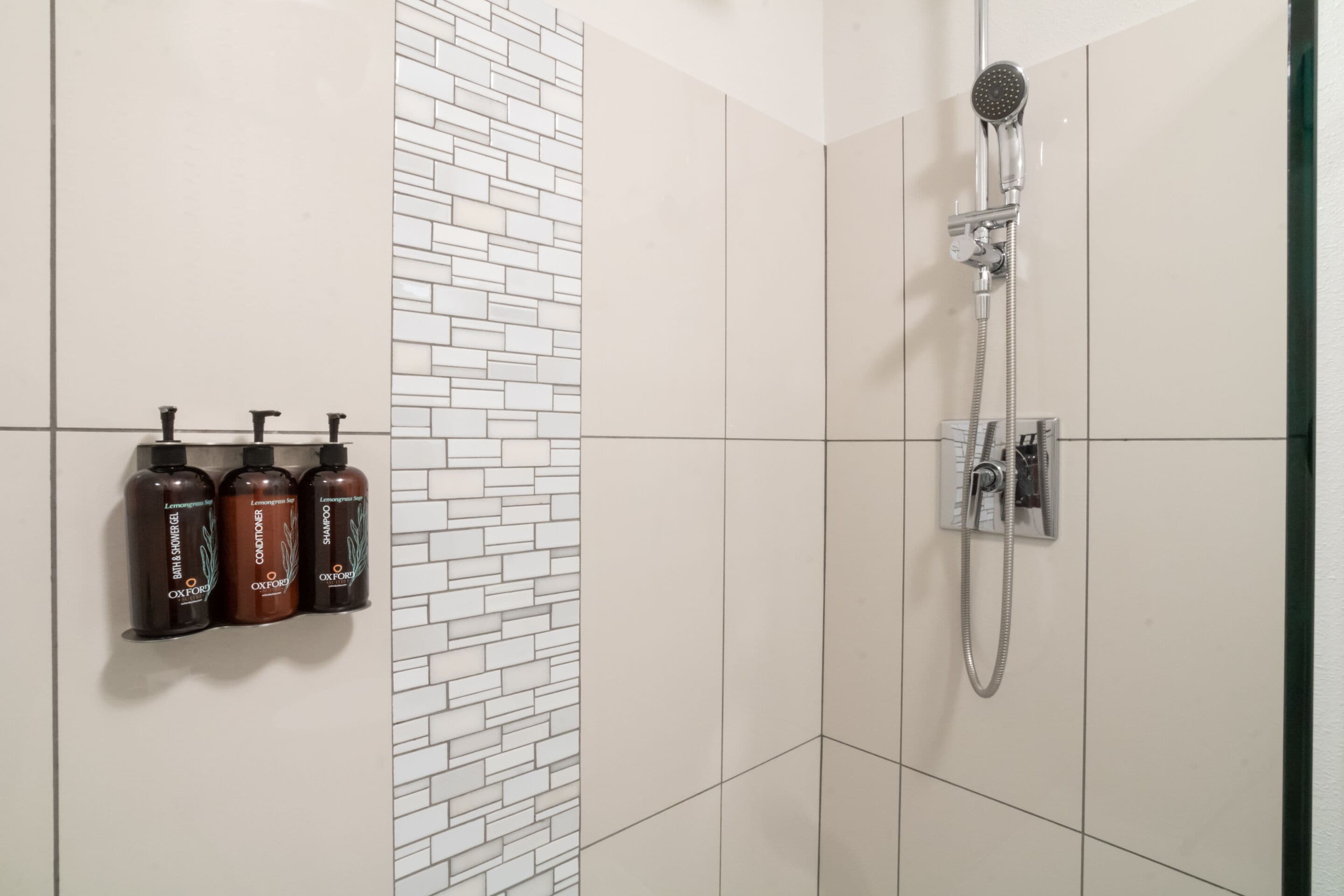 Une salle de bain avec une douche, trois bouteilles de shampoing et un mur carrelé.