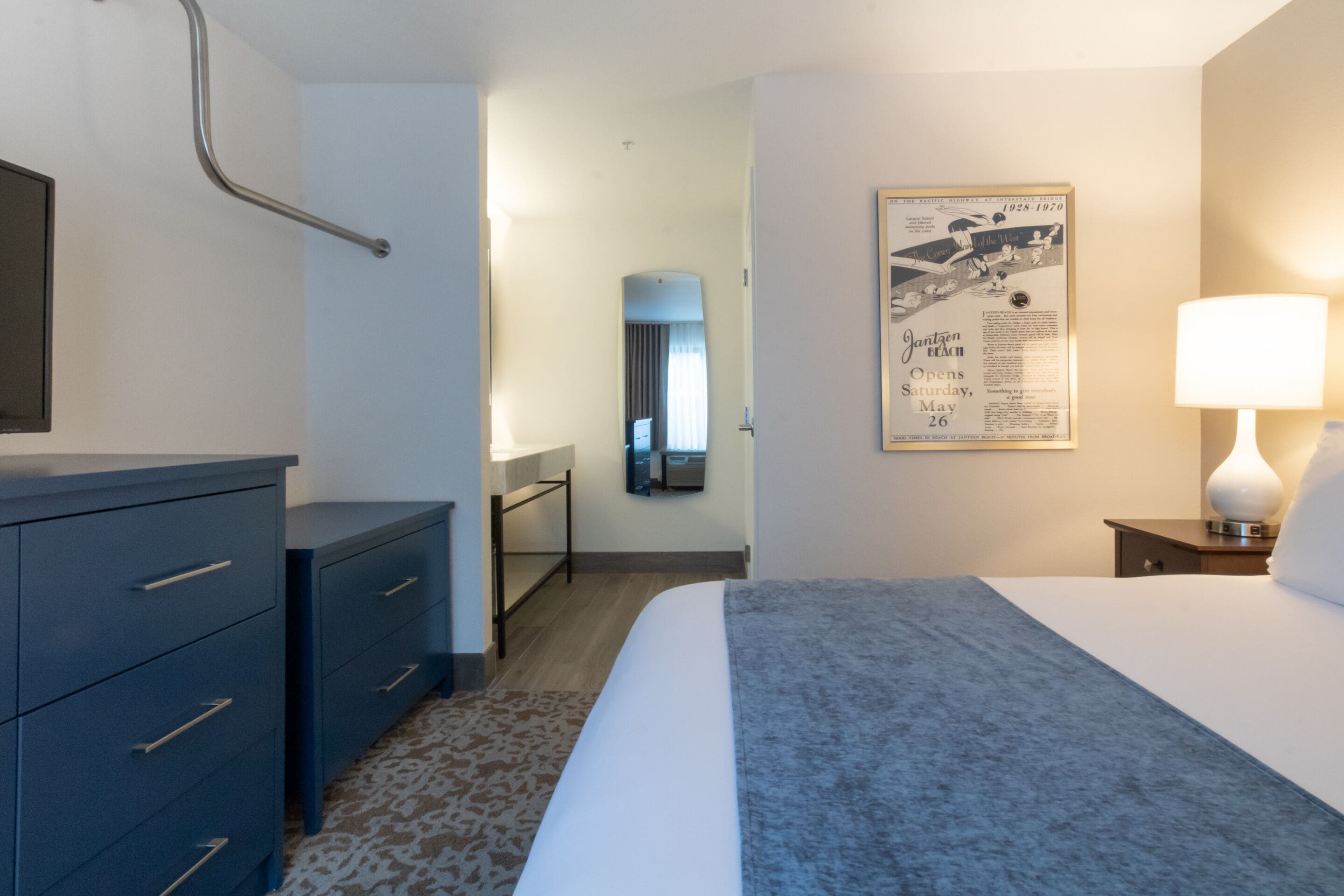 Habitación de hotel con cama, tocador, espejo, mesita de noche con lámpara y un cartel en la pared.