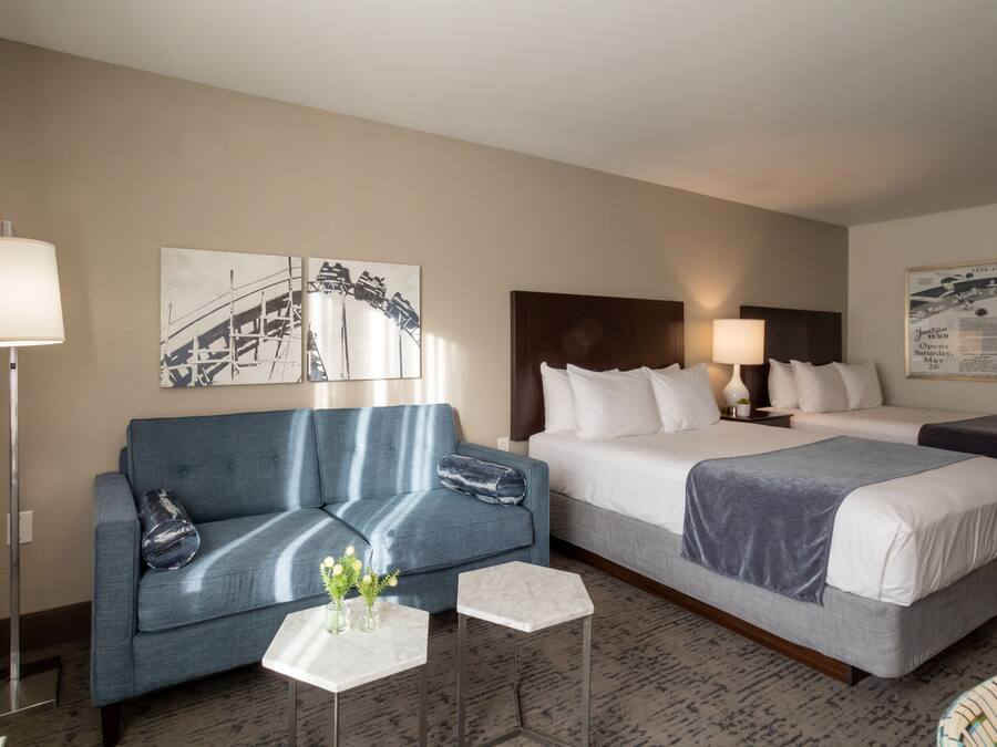 A hotel room with a blue sofa, a bed with a grey bedspread, a small table with a vase of flowers, and a lamp.