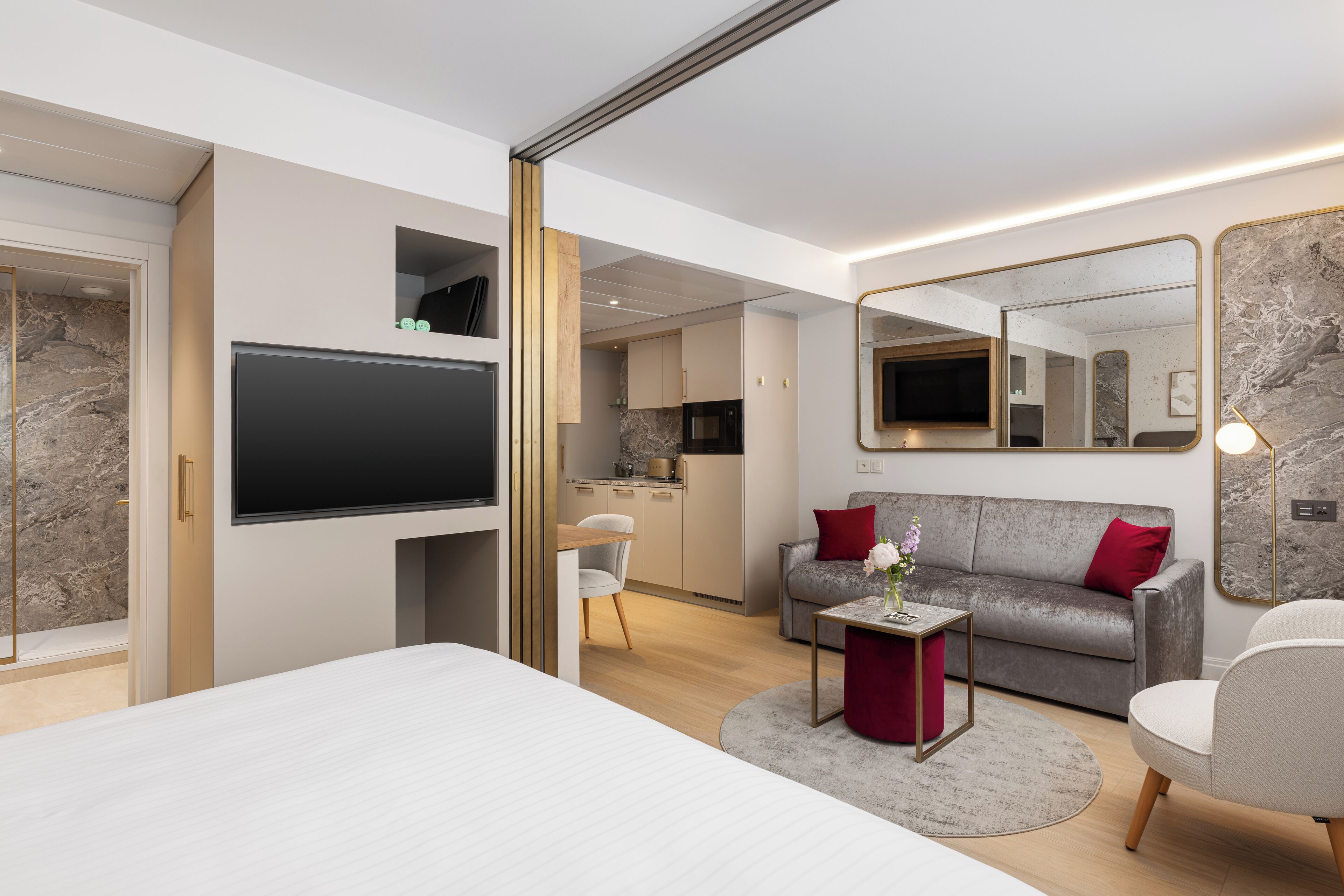 Una habitación de hotel moderna con una cama grande, un televisor montado en la pared, un sofá, una pequeña mesa con un jarrón, y una cocina visible a través de una puerta abierta.