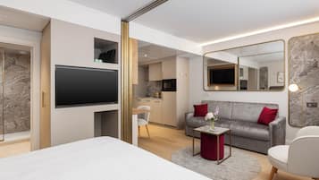Una habitación de hotel moderna con una cama grande, un televisor montado en la pared, un sofá, una pequeña mesa con un jarrón, y una cocina visible a través de una puerta abierta.
