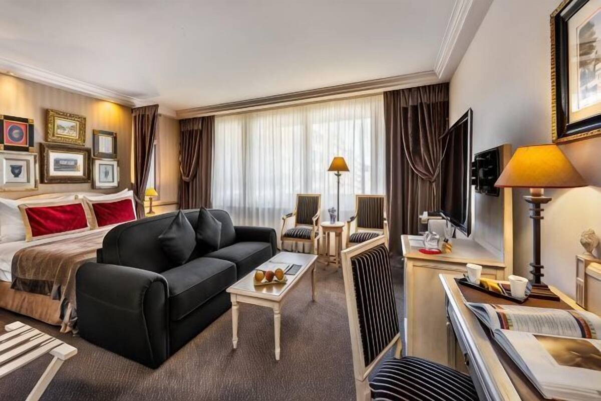 Hotellihuone, jossa on sänky, musta sohva, sohvapöytä, nojatuoli, televisio ja suuri ikkuna, jossa on verhot.
