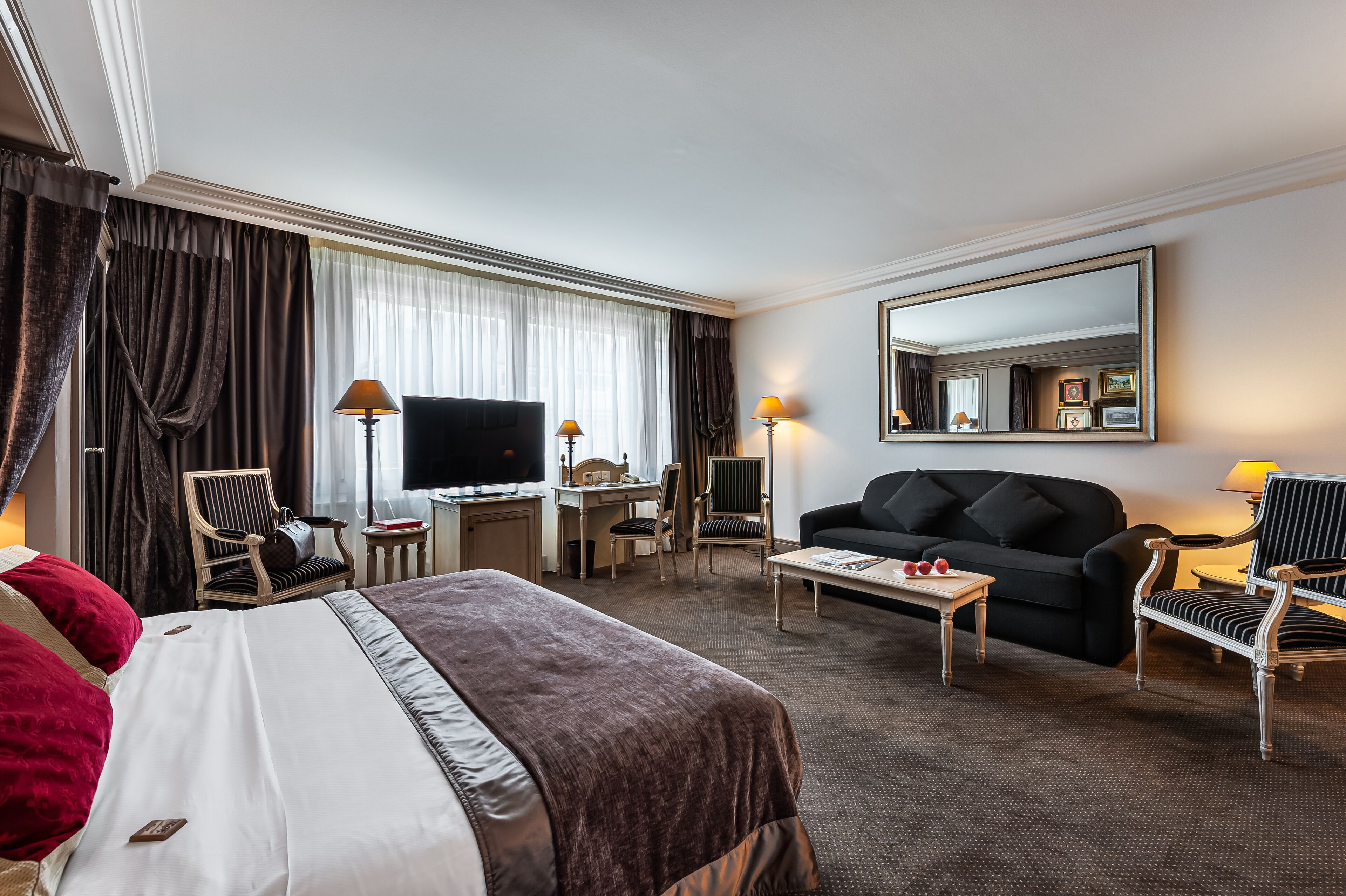 A hotel room with a large bed, a desk with a computer, a TV, a sofa, and two chairs.