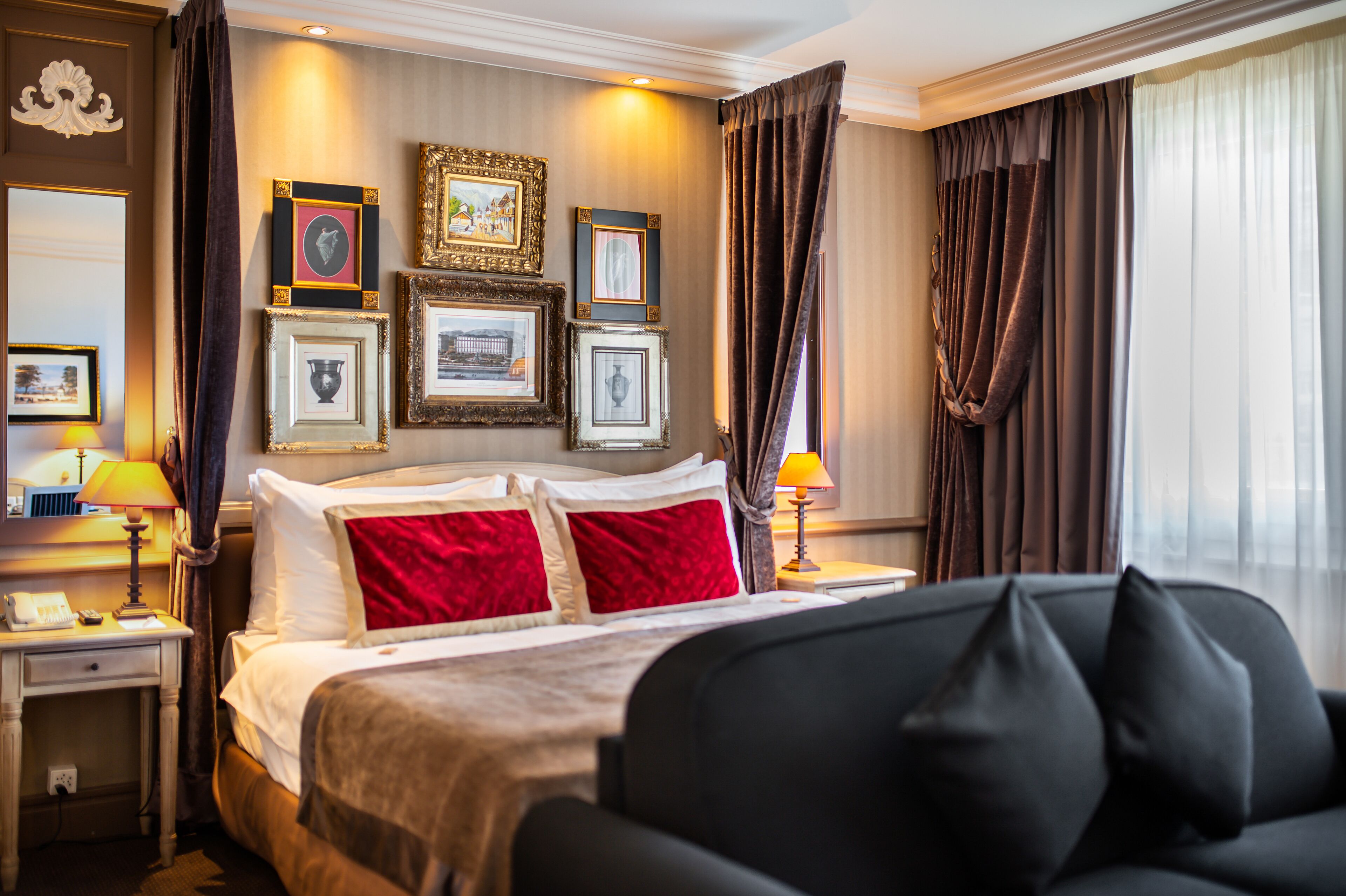 A hotel room with a large bed, two bedside tables, a nightstand, and a sofa. There are framed pictures on the wall and a window with curtains.