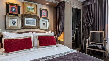 A hotel room with a large bed, red and white pillows, a framed picture collection on the wall, a chair, and dark curtains.