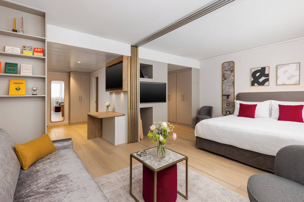 Moderni hotellihuone, jossa on suuri sänky, sohva, televisio ja pieni pöytä, jonka päällä on maljakko täynnä kukkia.