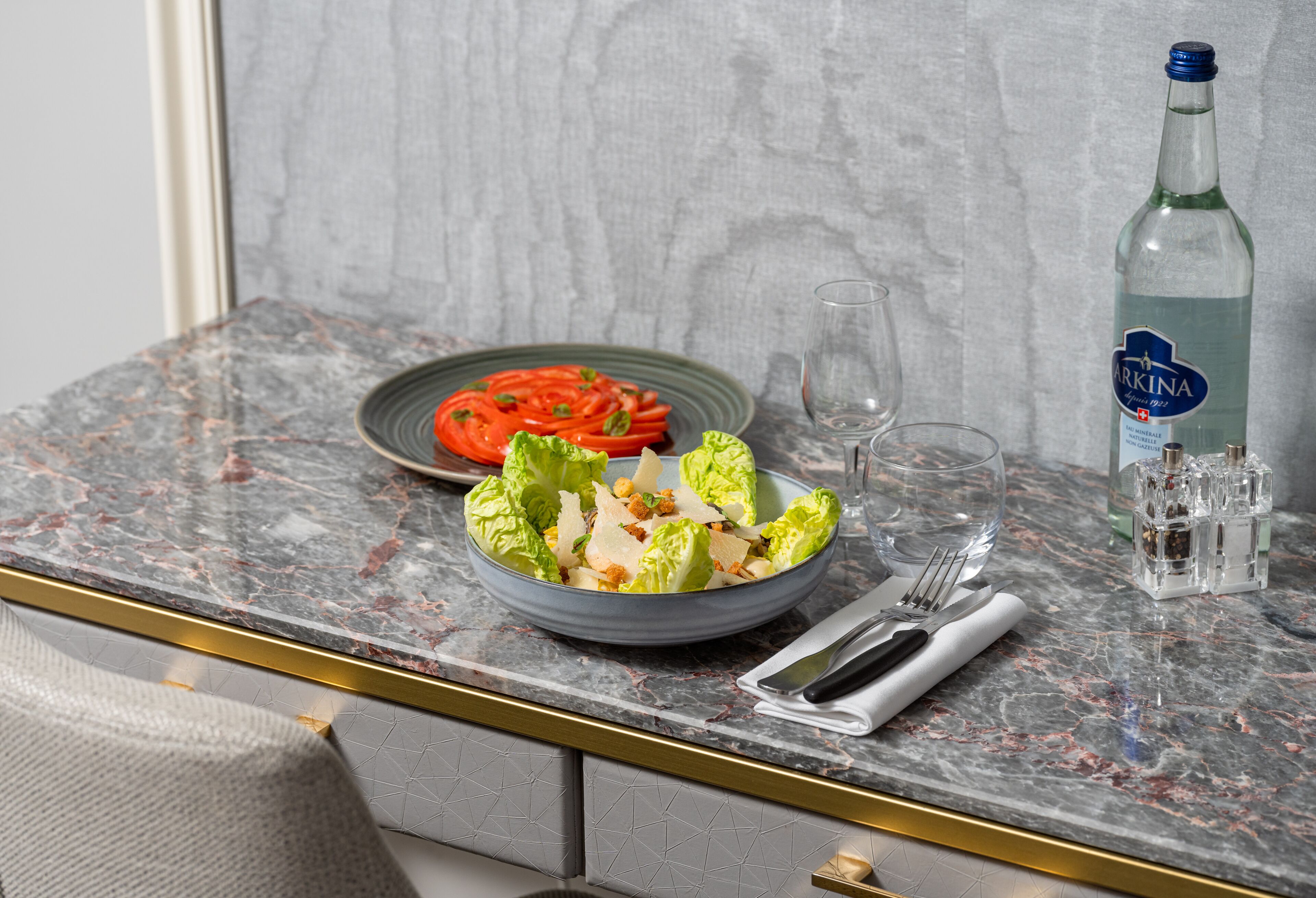 A marble countertop with a salad bowl, a plate of vegetables, a bottle of water, and a glass.