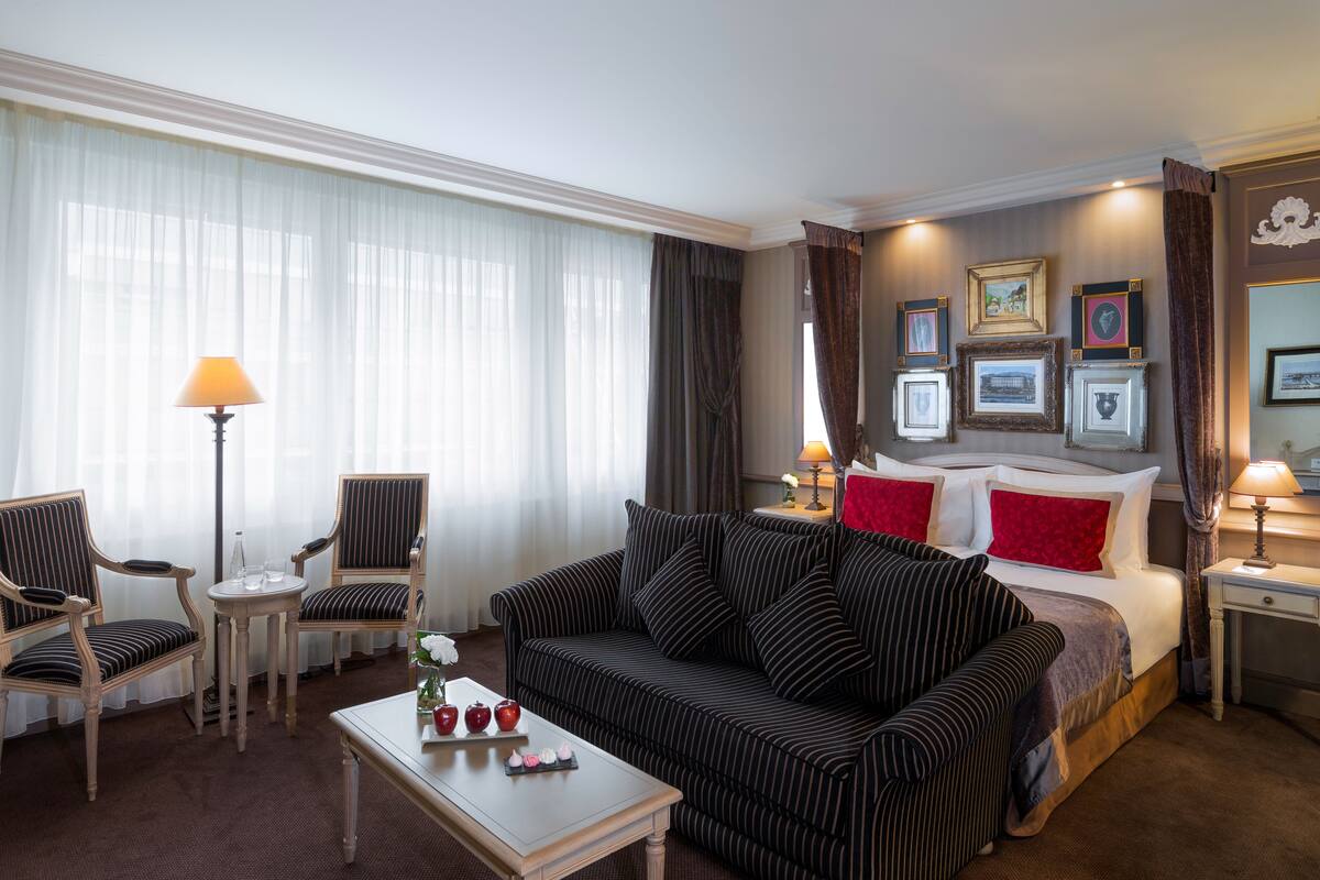 Hotellihuone, jossa on suuri sänky, sohva, kaksi nojatuolia, pieni pöytä, maljakko ja hedelmiä, sekä lamppu.