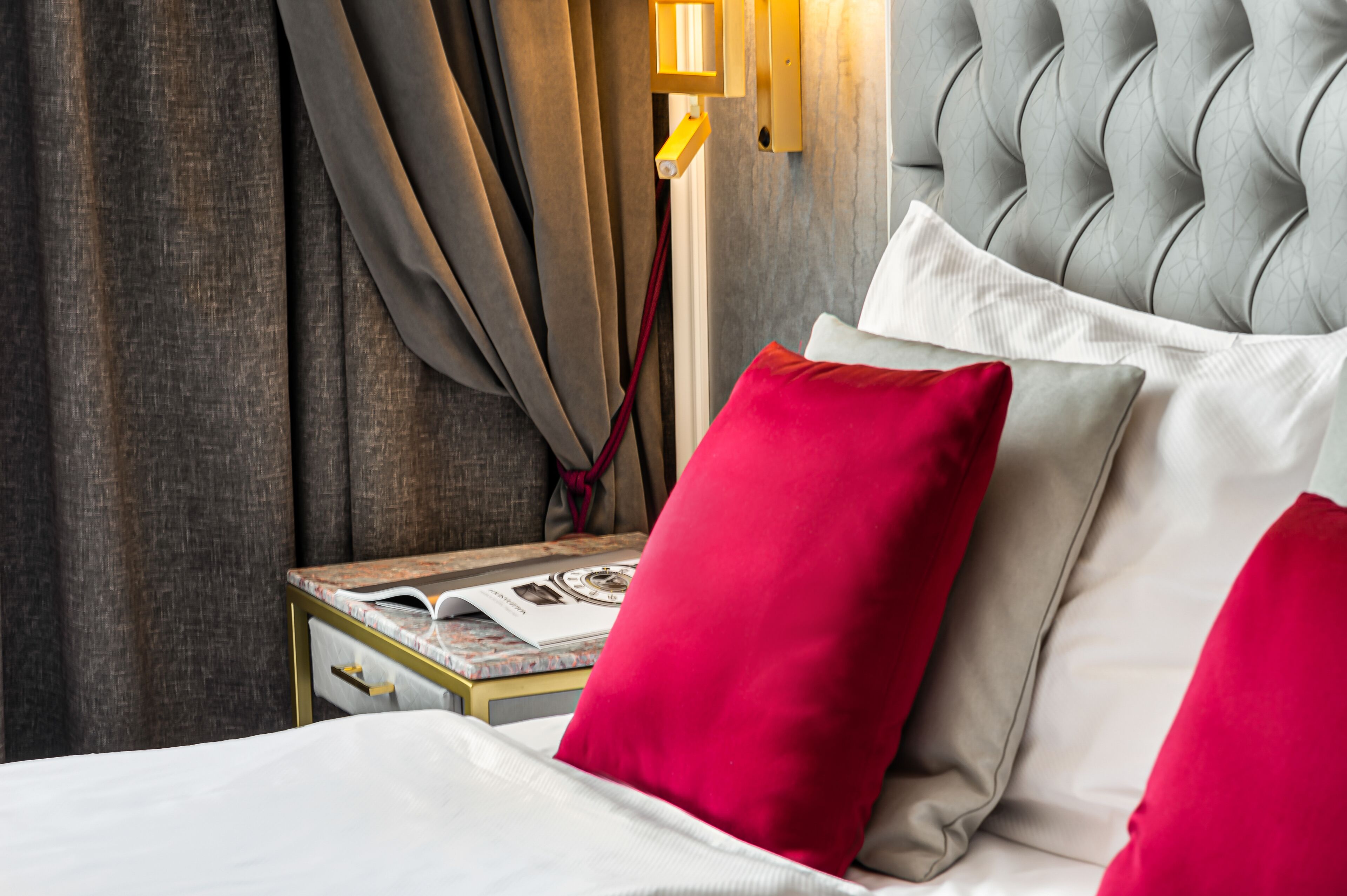 Une chambre d’hôtel avec une tête de lit capitonnée, une table de chevet sur laquelle repose un livre, et des oreillers rouges et blancs.