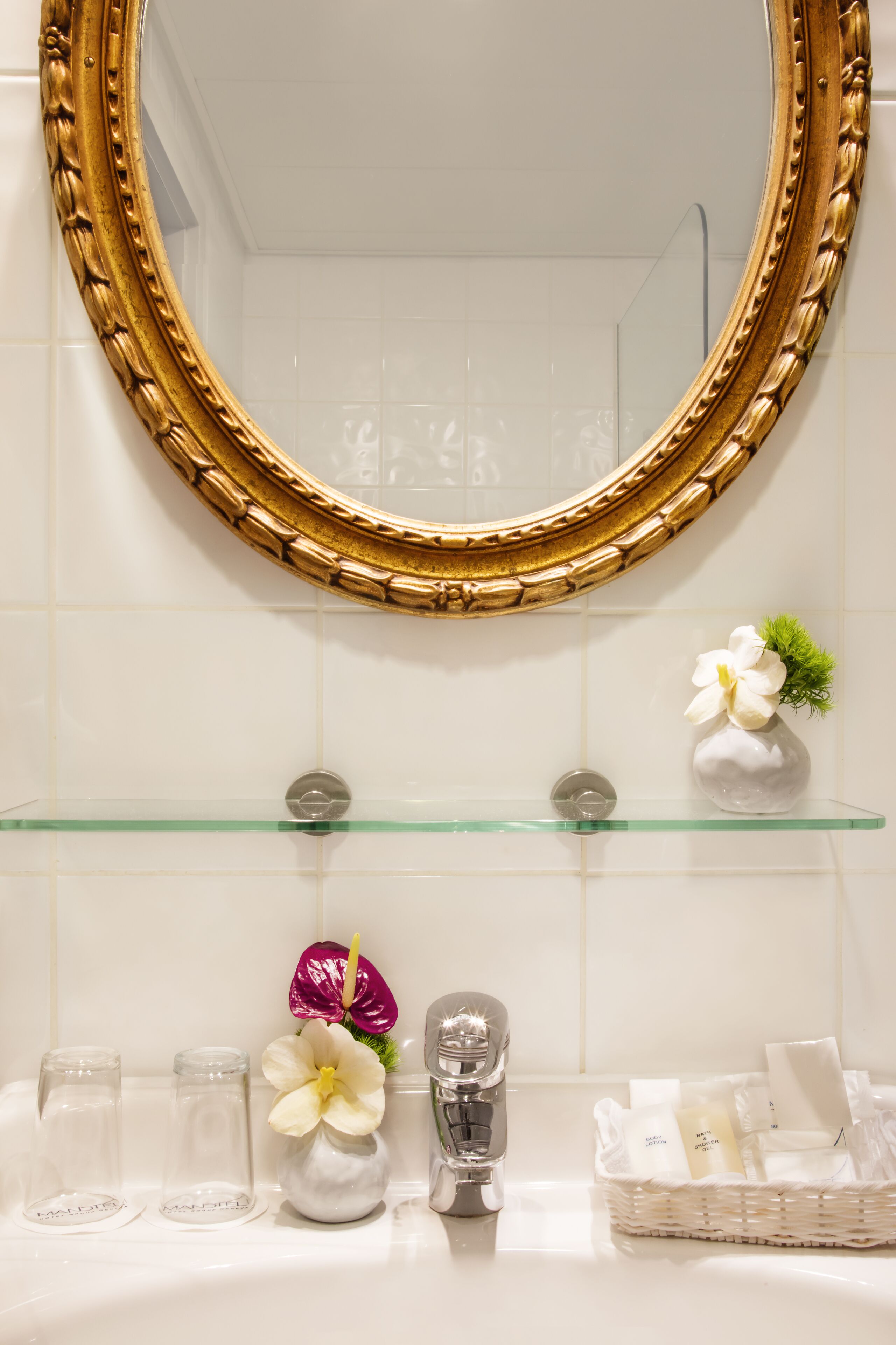 Un baño con un espejo de marco dorado, un estante de vidrio con artículos de aseo y un lavabo blanco.