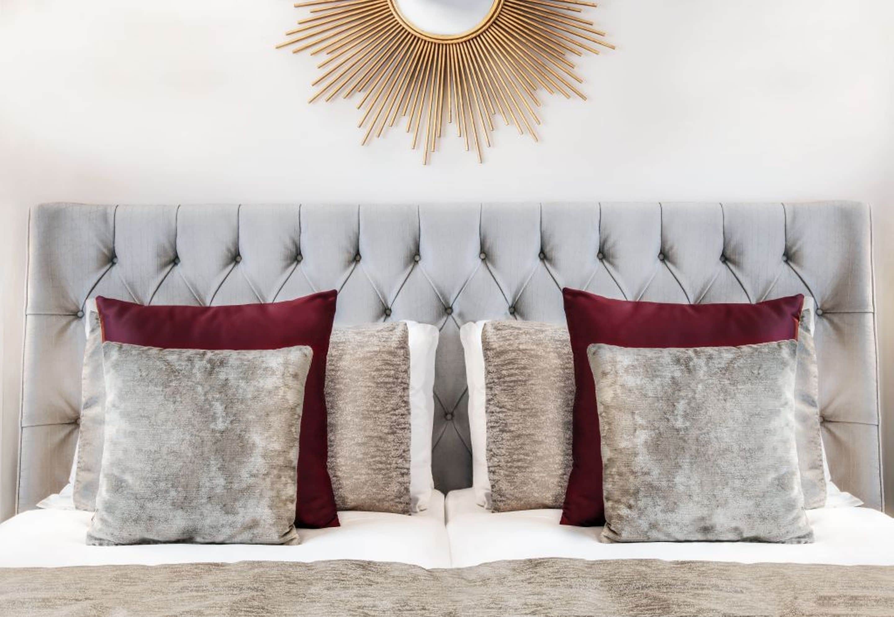 Una cama con cabecera capitonada, dos almohadas y un espejo con forma de sol dorado en la parte superior.