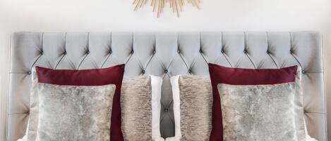 Una cama con cabecera capitonada, dos almohadas y un espejo con forma de sol dorado en la parte superior.
