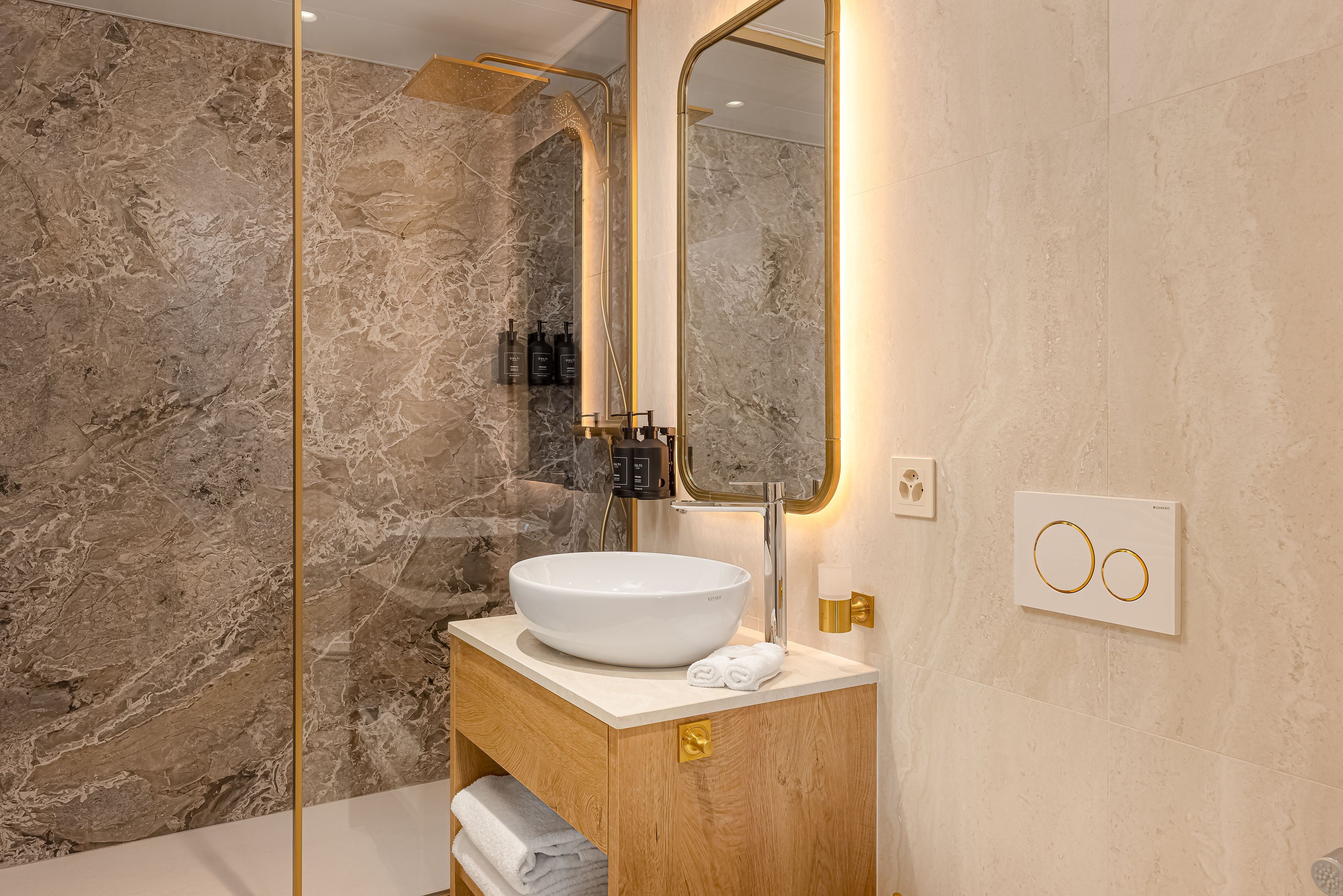 Un baño con pared de mármol, un lavabo blanco y un espejo con marco dorado.