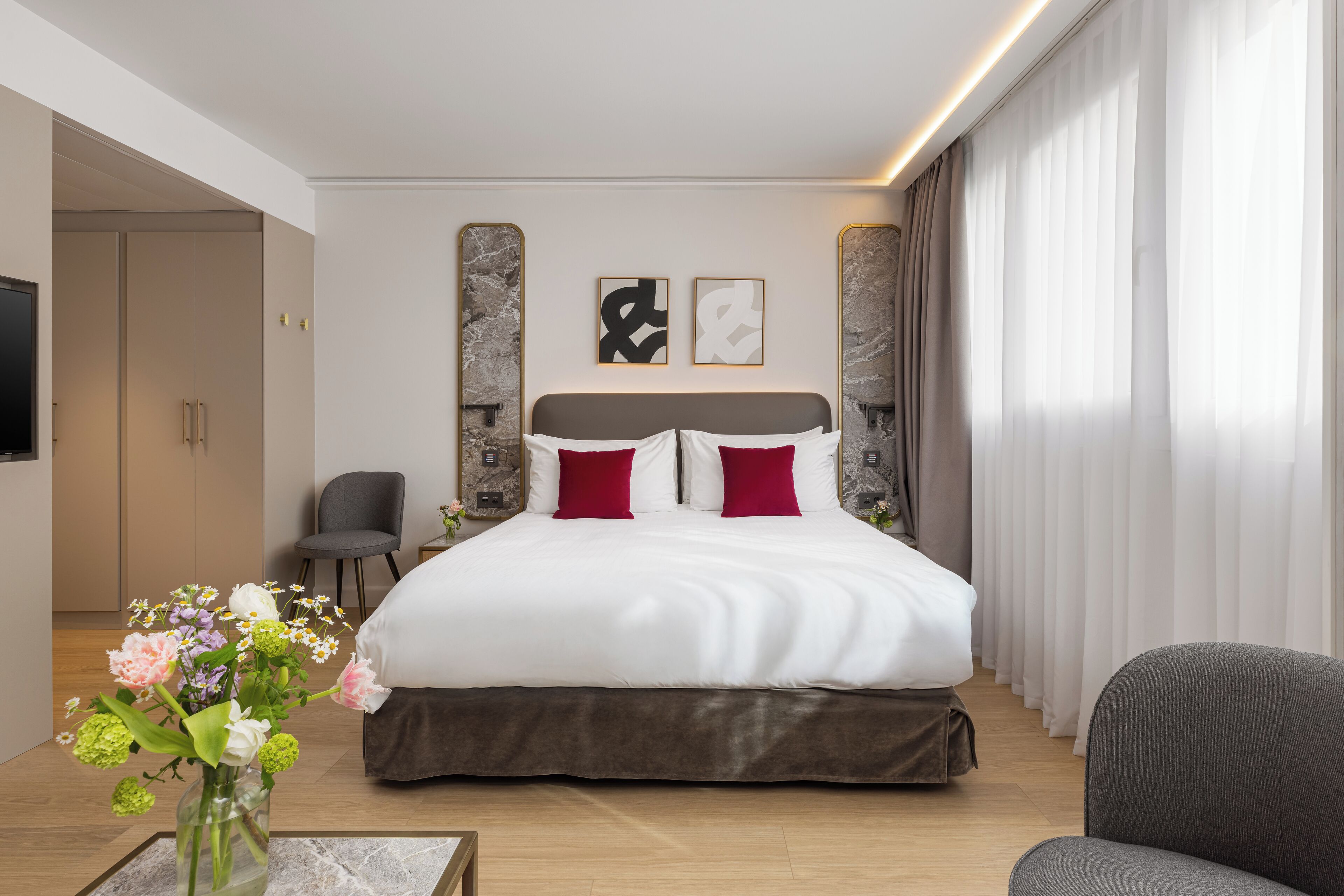 Une chambre d’hôtel moderne avec un grand lit, un téléviseur à écran plat, un vase de fleurs et un fauteuil gris.