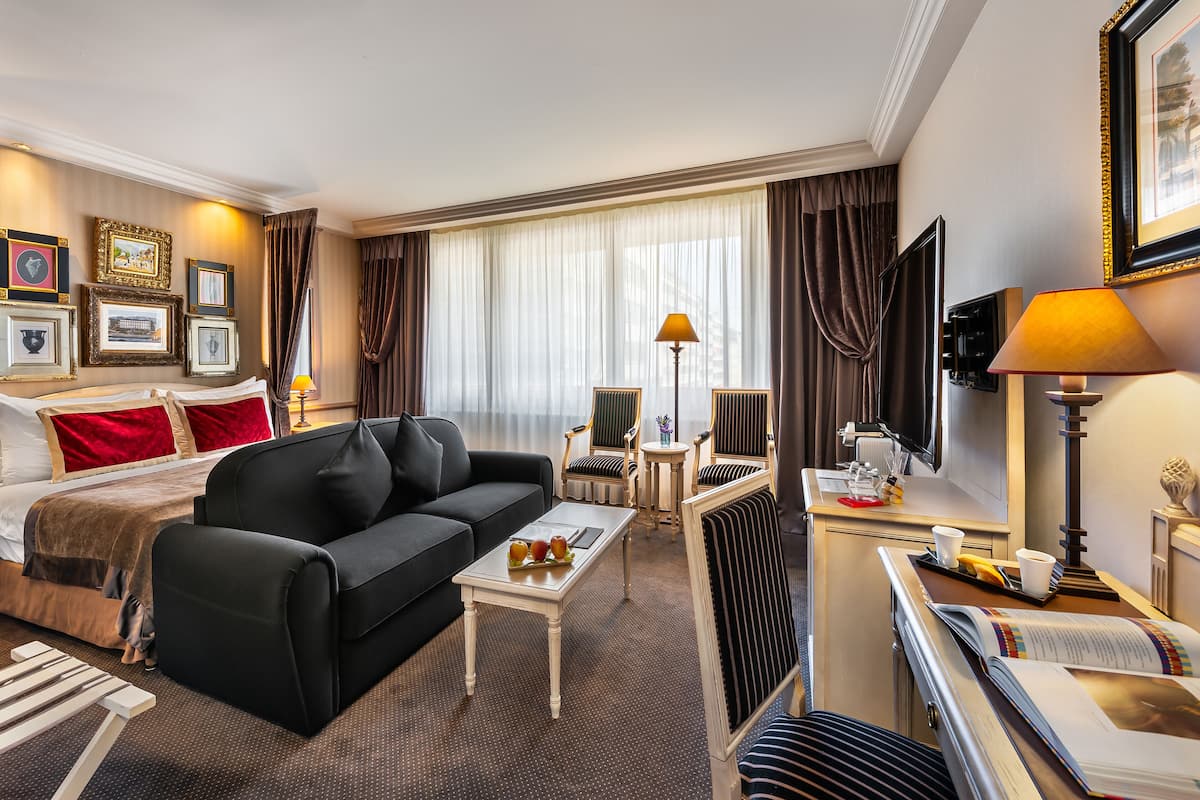 Hotellihuone, jossa on sänky, musta sohva, ruokapöytä, tuolit, televisio ja seinällä olevia tauluja.
