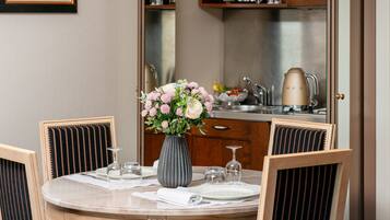 Un comedor con una mesa redonda preparada para dos personas, un florero con flores y una cocina al fondo.
