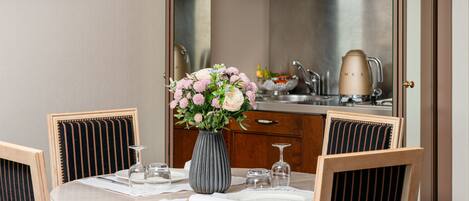 Un comedor con una mesa redonda preparada para dos personas, un florero con flores y una cocina al fondo.