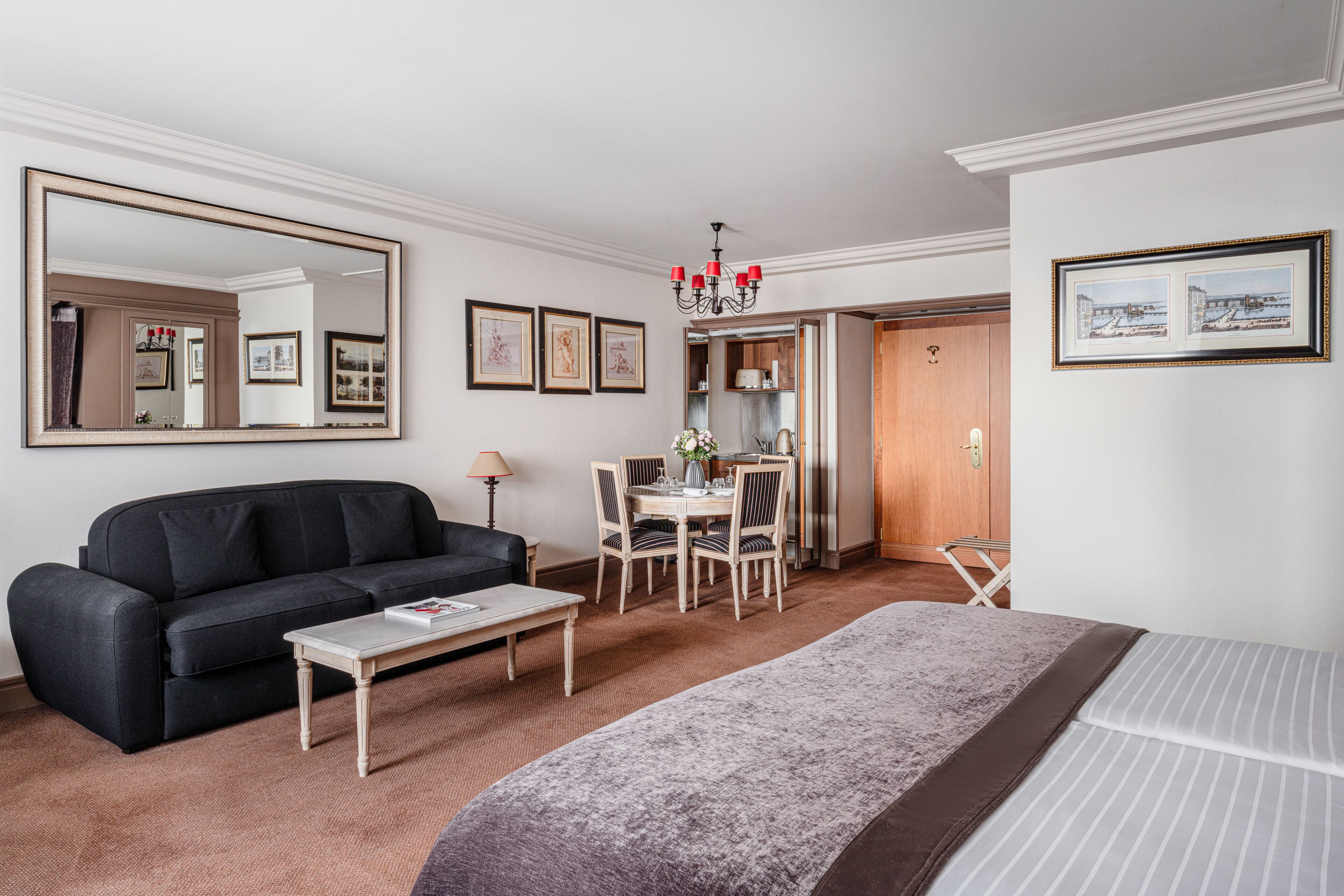 A hotel room with a bed, a sofa, a dining table with chairs, a coffee table, a large mirror, and a chandelier.