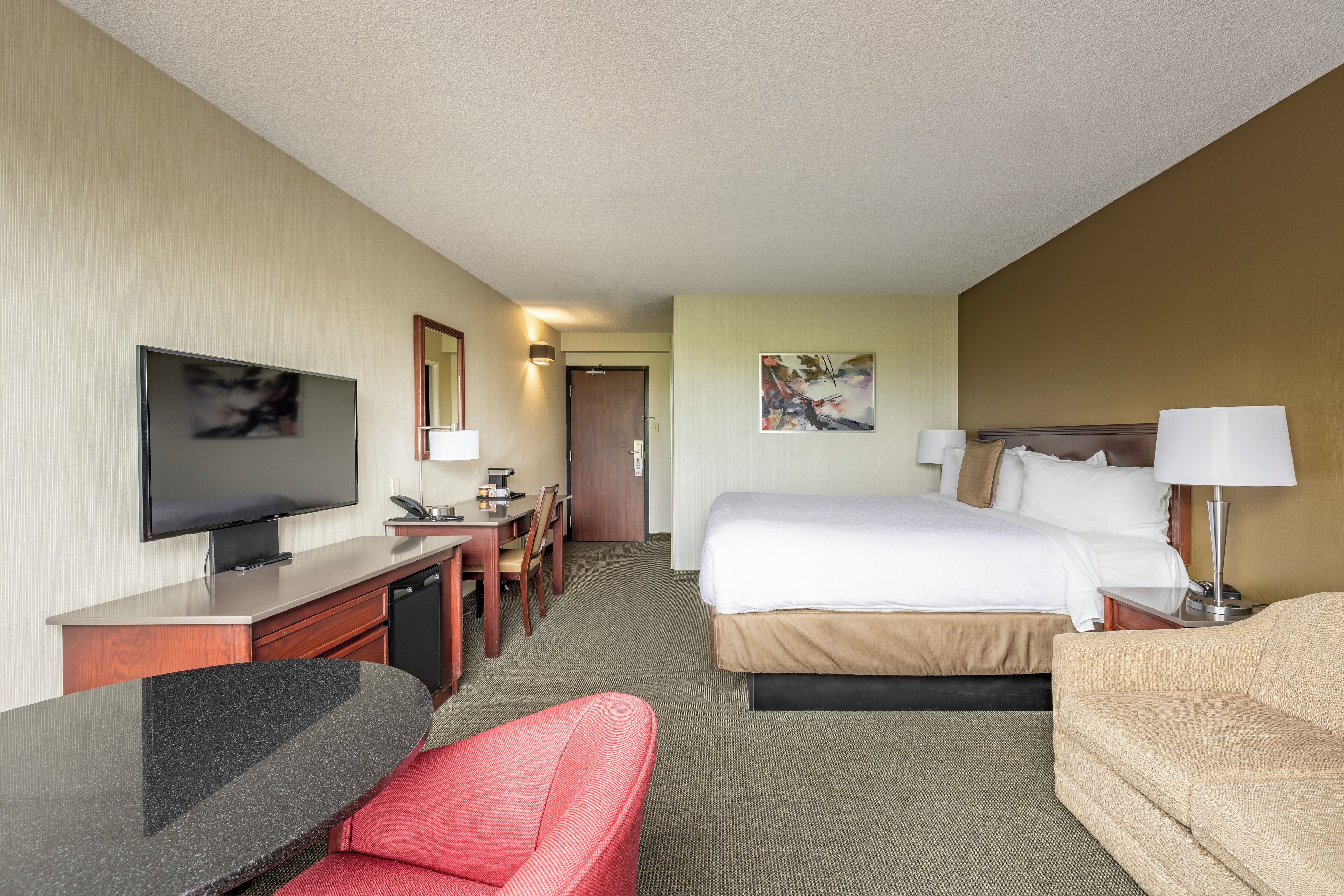 A hotel room with a bed, a desk with a television, a chair, a sofa, and a painting on the wall.