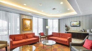 Un cuarto de hotel con dos sofás rojos, una mesa de centro de vidrio, un sillón color beige, una mesita con una lámpara y un televisor en la pared.