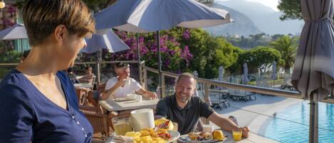 Comedor con asientos exteriores cerca de una piscina, con sombrillas y vistas a montañas.