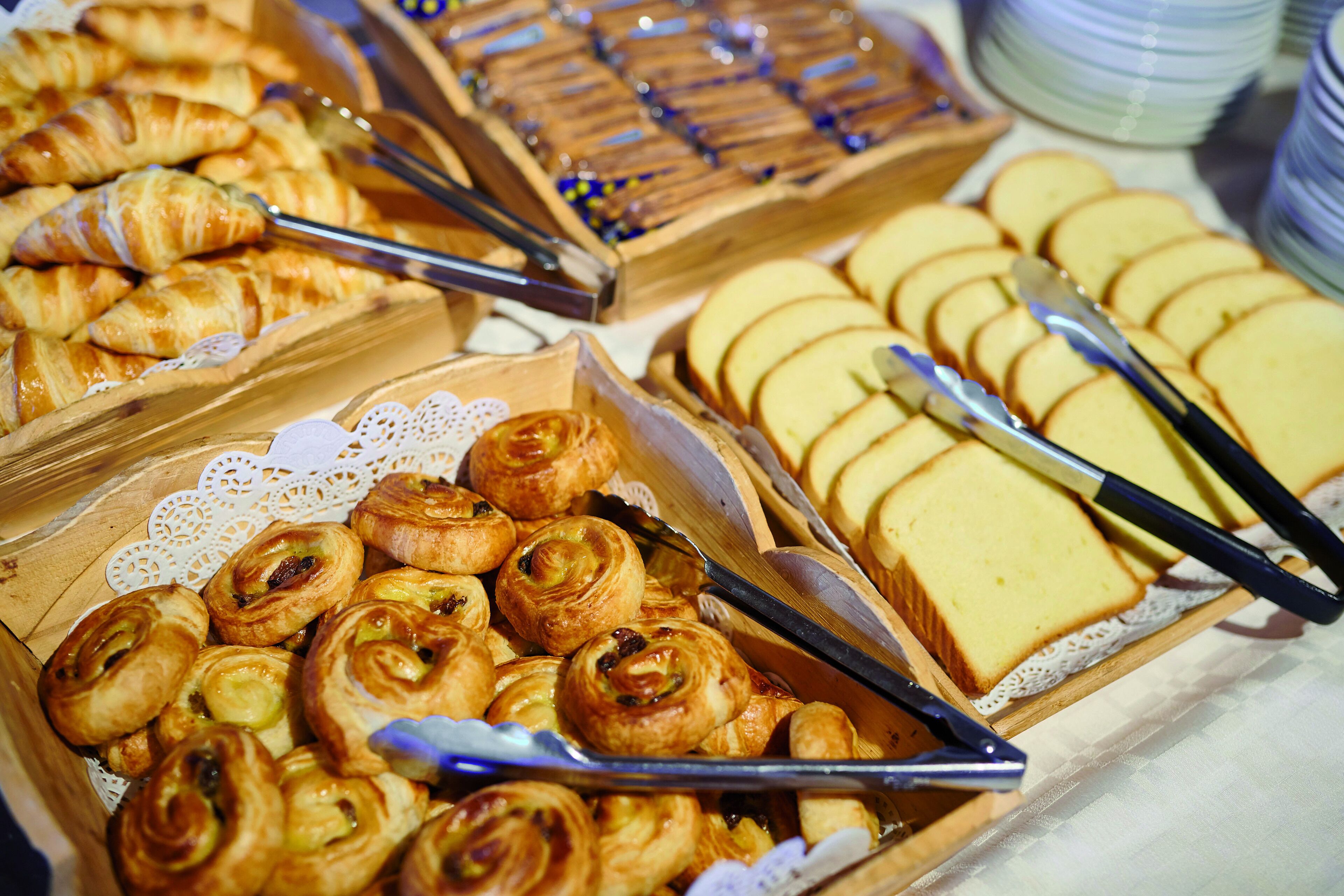 A variety of pastries and bread displayed in wooden trays with tongs.