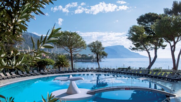 A pool with a unique sculpture centerpiece, surrounded by lounge chairs and trees, with a mountain and sea in the background. - Hôtel Vacances Bleues Delcloy (Saint-Jean-Cap-Ferrat)