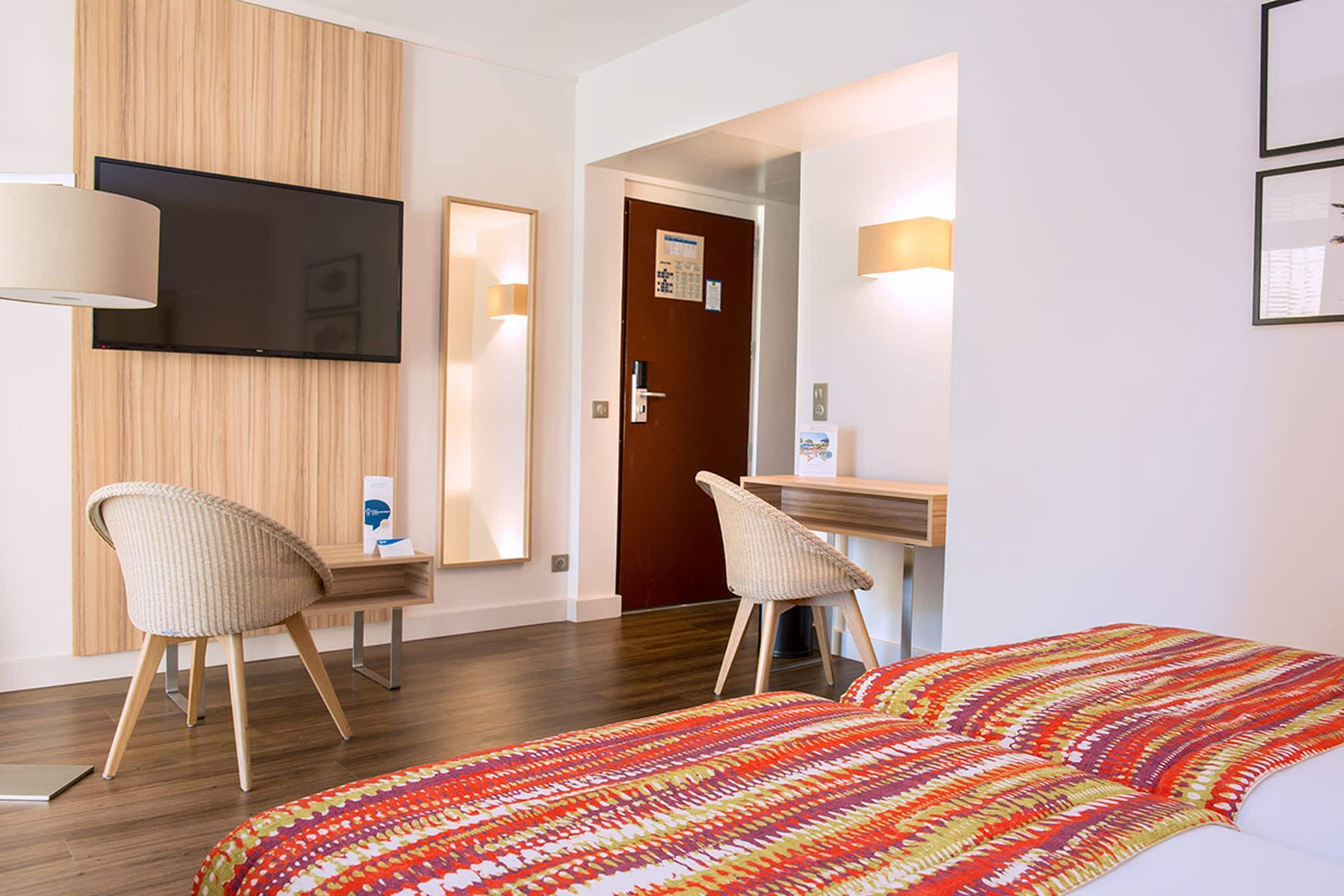 Habitación de hotel con cama, dos sillas, escritorio, televisor y puerta.