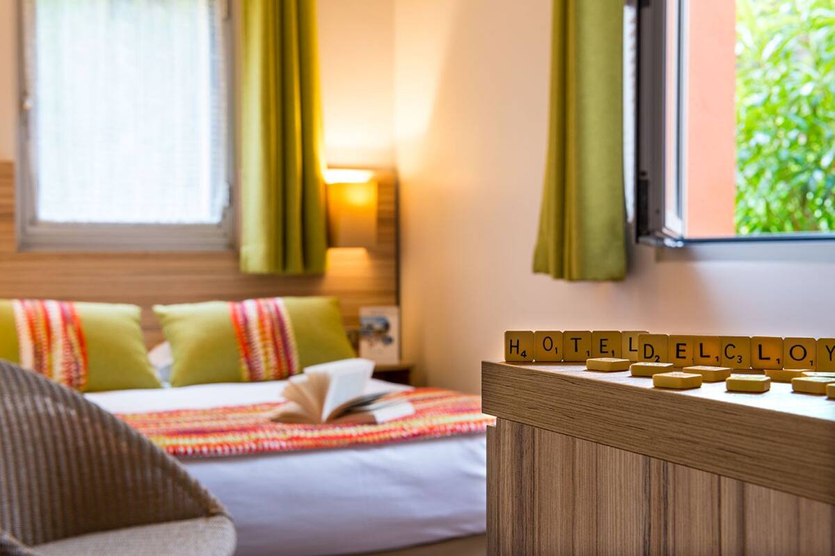 A hotel room with a bed, a bedside table with Scrabble tiles spelling "HOTEL," and a window with curtains.