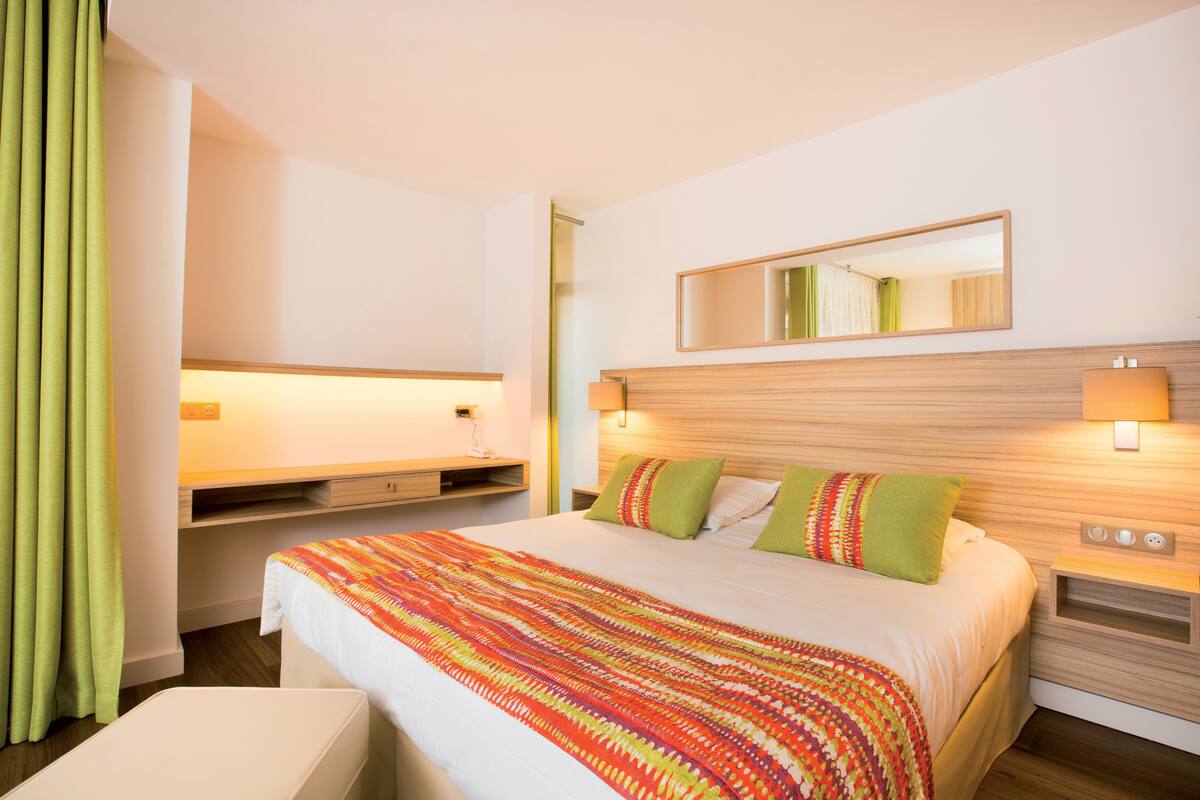 A hotel room with a large bed, a bedside table, a nightstand, a mirror, and a window with curtains.