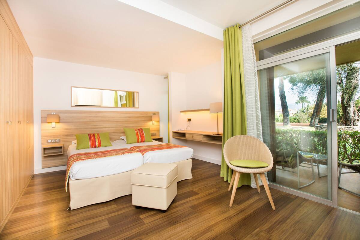 A modern hotel room with a wooden floor, a large bed with a green and orange striped pillow, a small table, a chair, and a sliding glass door leading to a balcony with greenery.