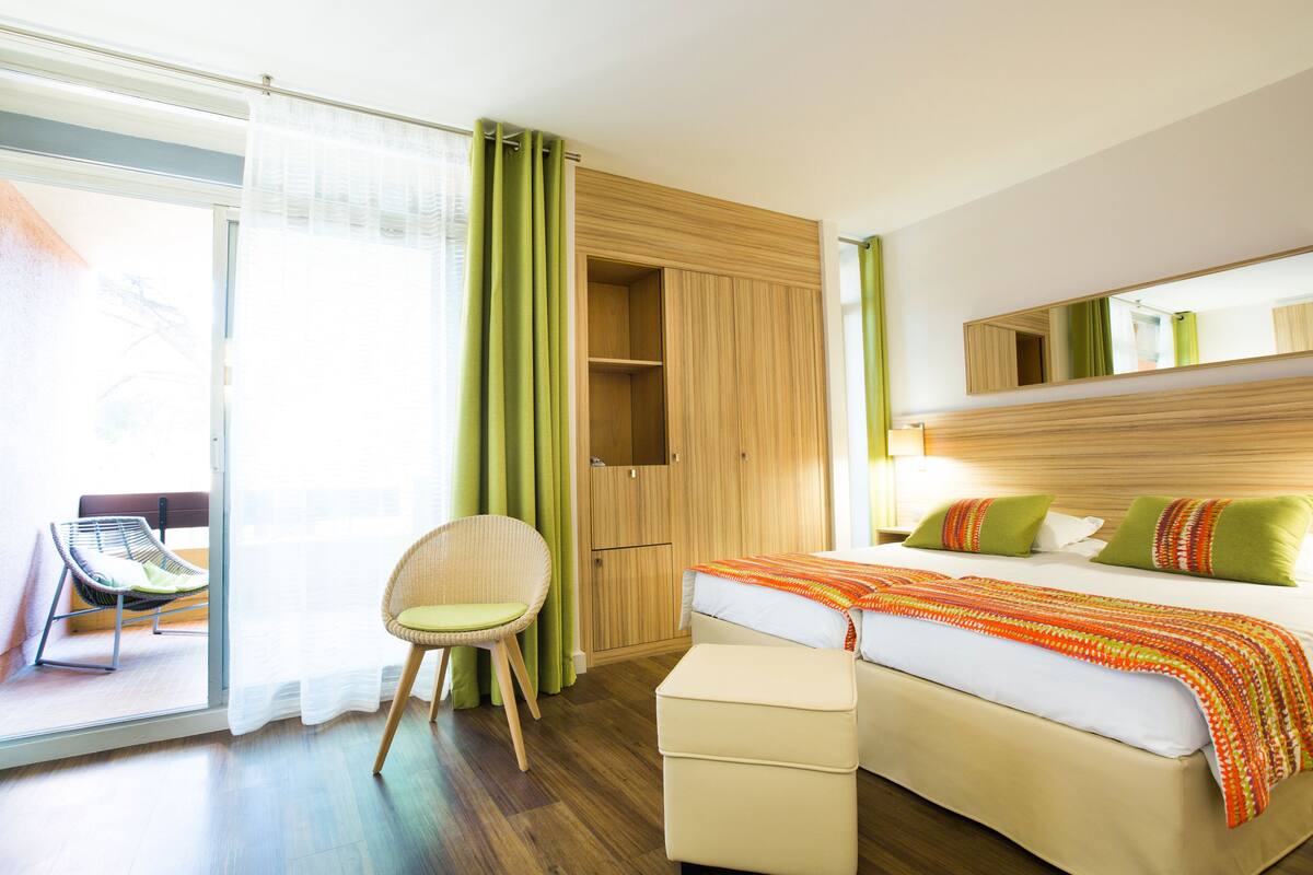 A hotel room with a bed, a chair, a wardrobe, and a sliding glass door leading to a balcony.