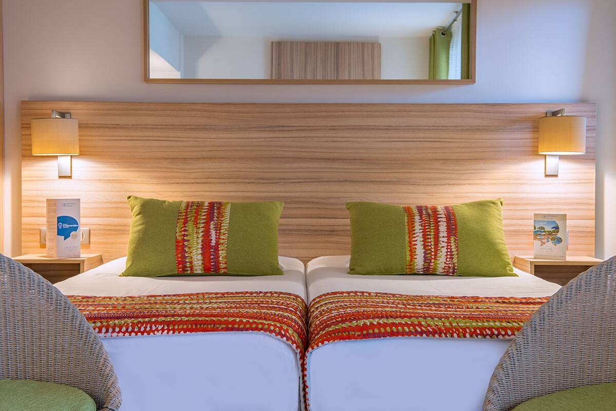 A hotel room with two beds, each with a green pillow and a patterned bedspread. There are two bedside lamps, a mirror above the beds, and a wooden headboard.