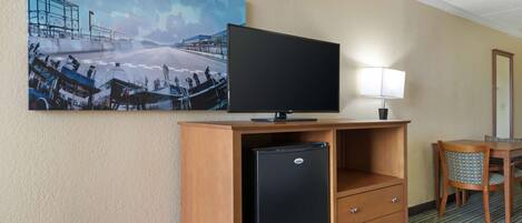 Habitación de hotel con televisión sobre un soporte de madera, un refrigerador pequeño, un escritorio con una silla y un cuadro en la pared.