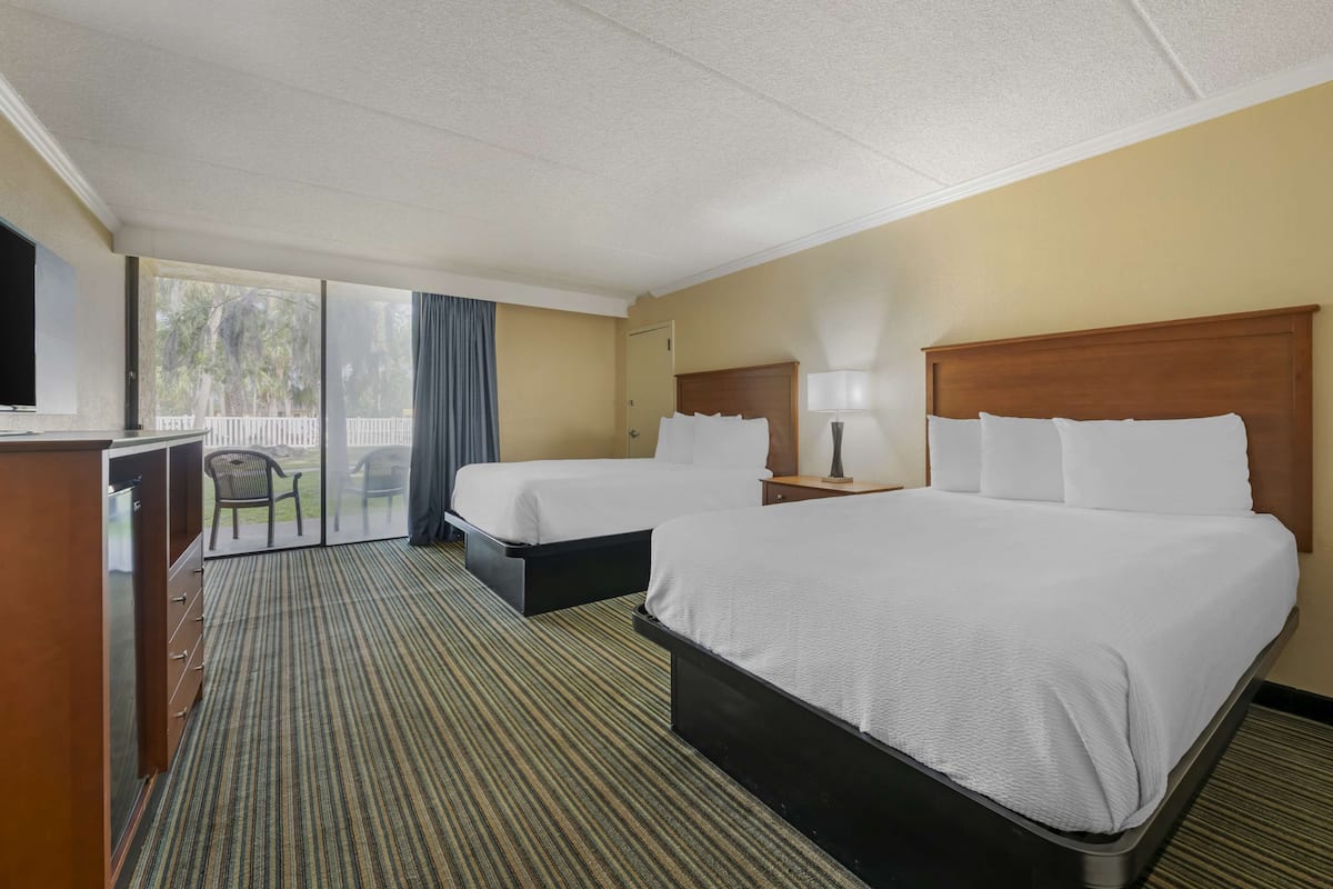 A hotel room with two beds, a desk with a TV, a chair, and a view of palm trees through the window.