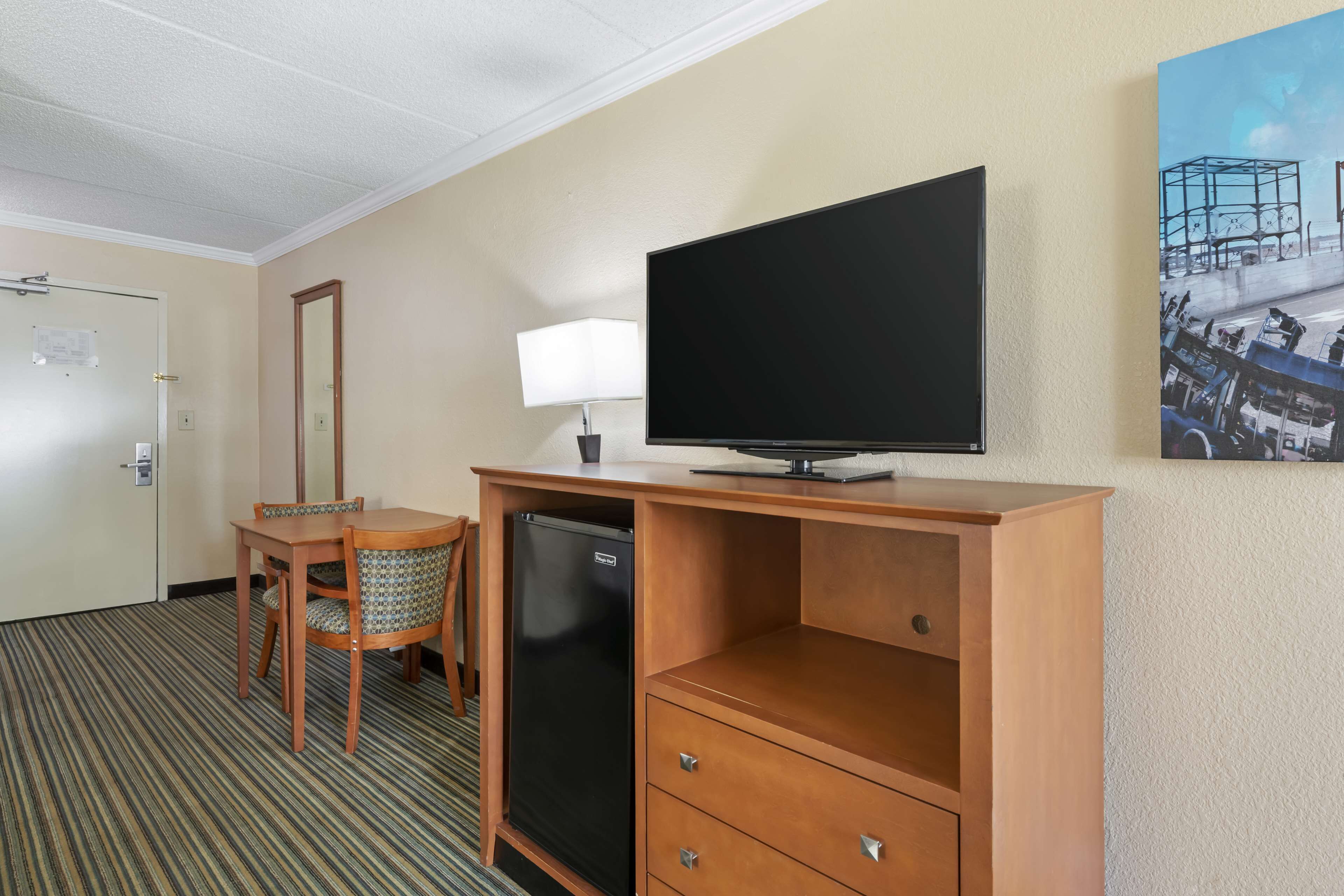 Habitación de hotel con escritorio de madera, televisor, refrigerador, lámpara y un cuadro en la pared.